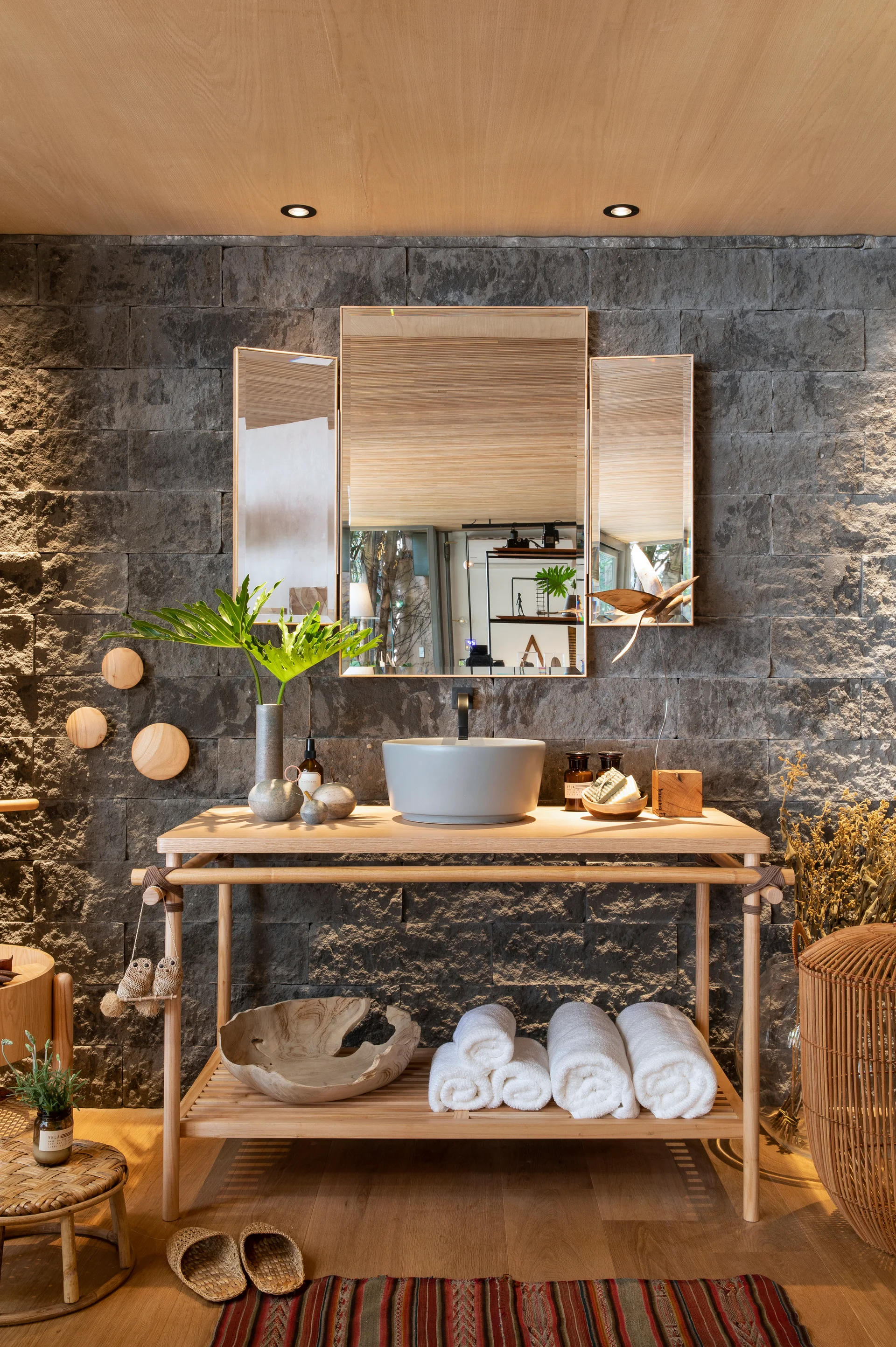A light wood console vanity with a grey vessel sink stands against a rough dark stone wall beneath a three-panel mirror.