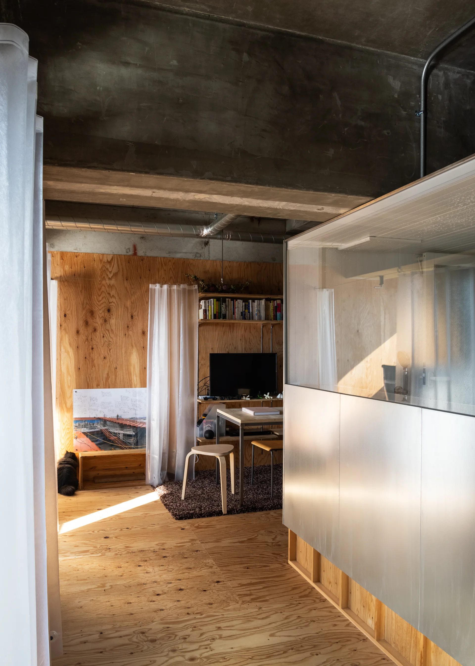 A modern apartment interior features plywood walls and floors, an exposed concrete ceiling, and a metal partition next to a small wooden dining nook.