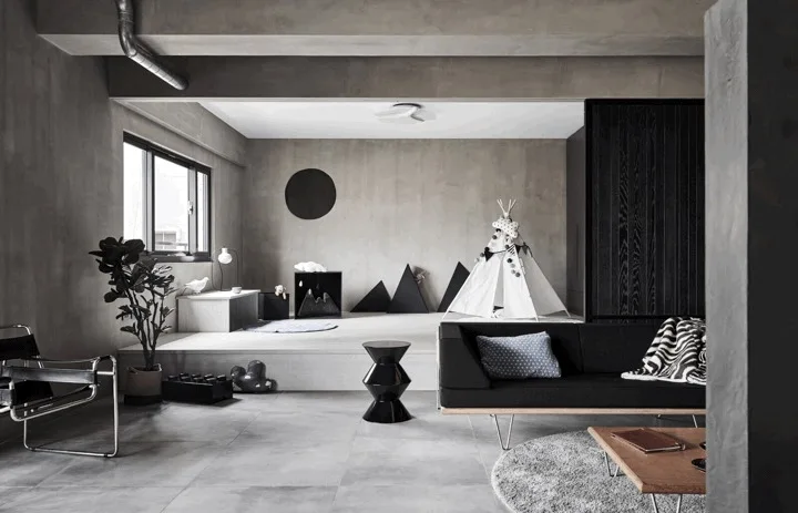 A minimalist grey concrete living room features a black sofa, a geometric side table, and a raised platform with a children's play teepee.