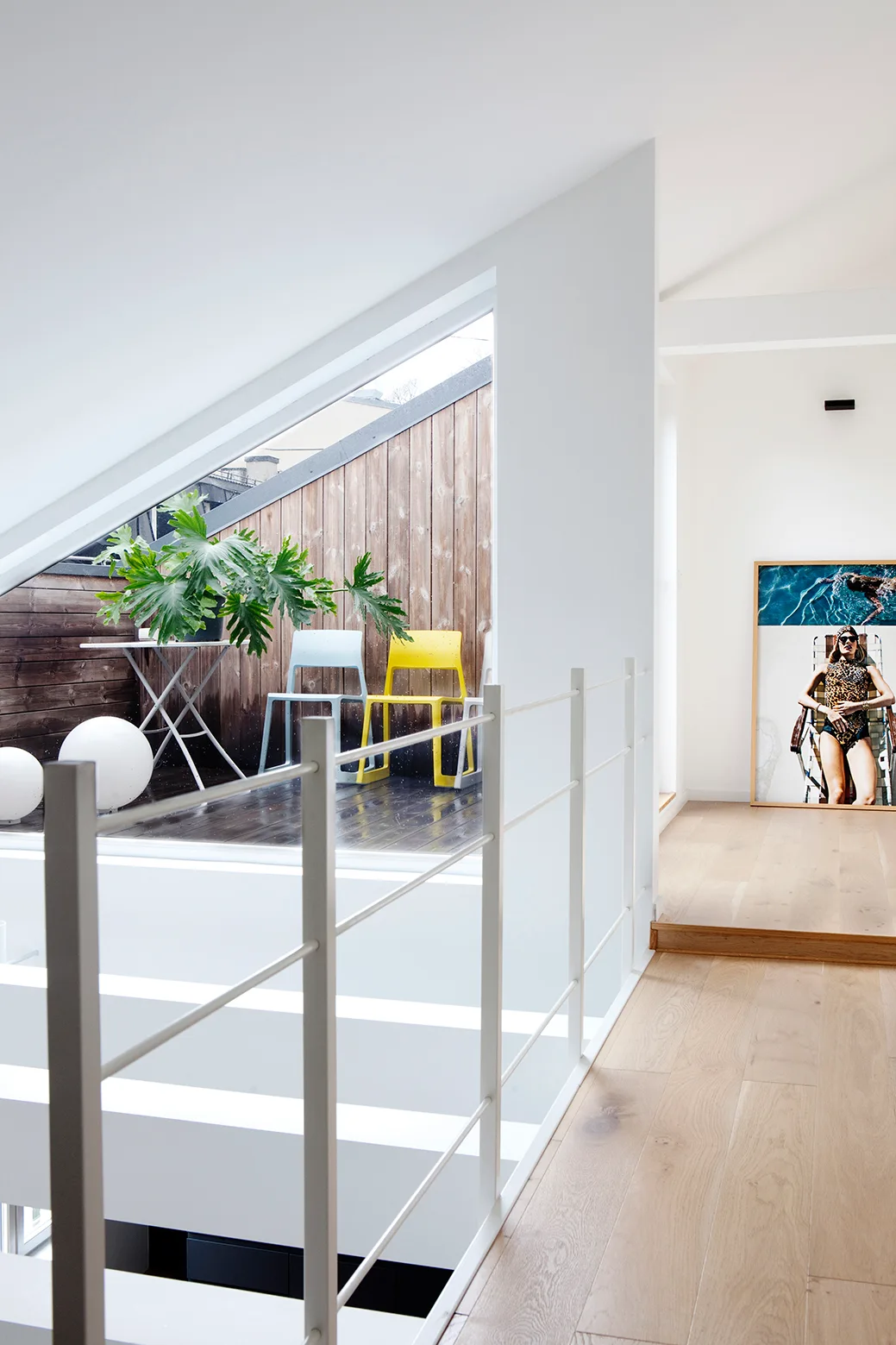A minimalist hallway landing with a white railing and light hardwood flooring overlooks a wood-clad balcony with two colorful chairs.