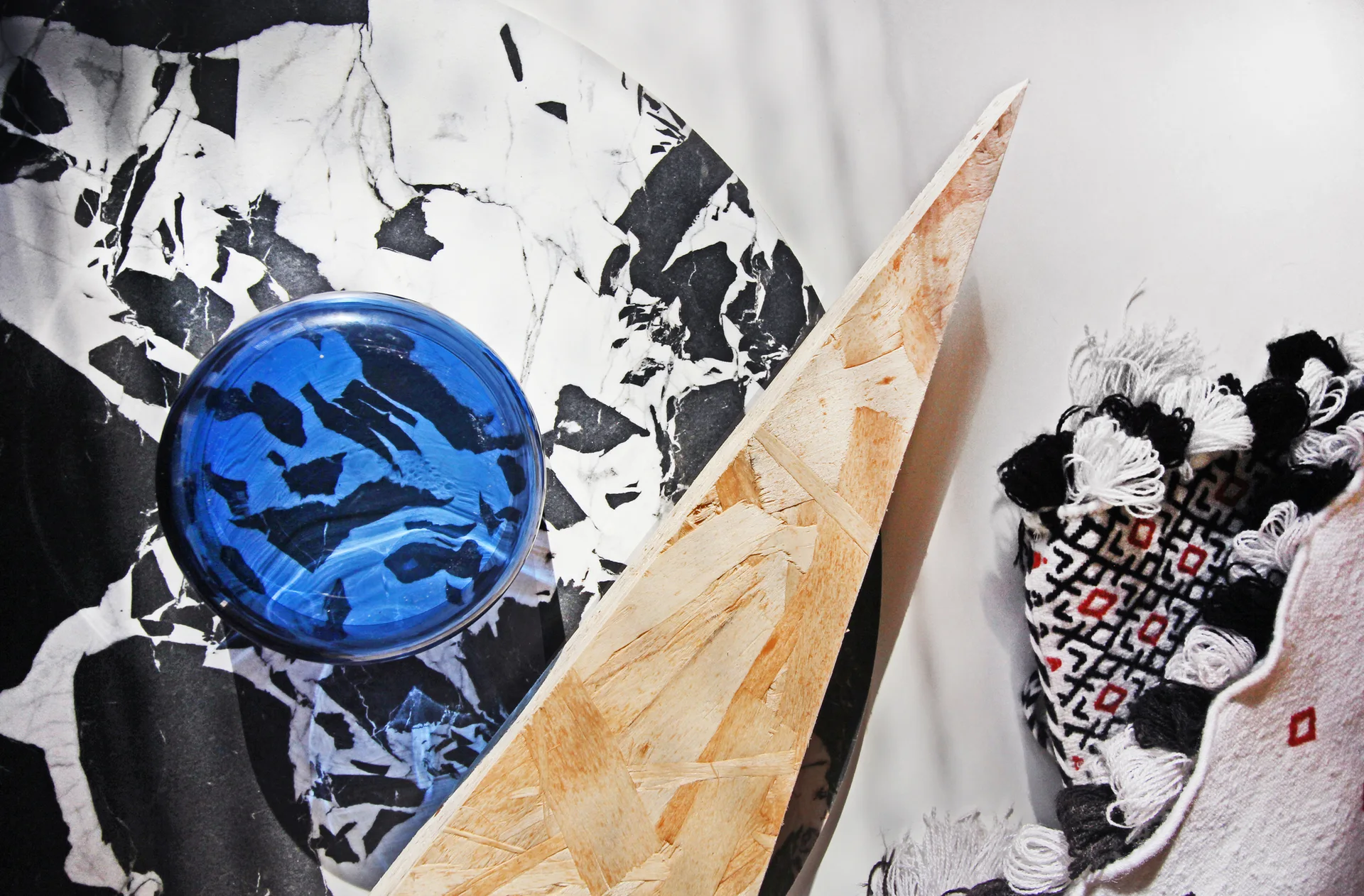 A flat lay of design material samples featuring a black and white marbled tray, a blue glass bowl, a triangular wood wedge, and a tasseled fabric swatch.