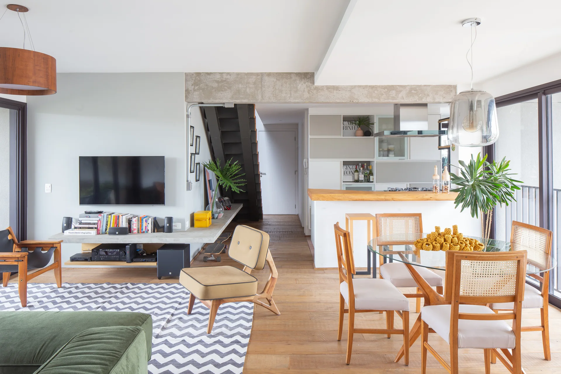 An open-concept living and dining area features a glass-top table with cane chairs, a concrete ceiling beam, and a chevron patterned rug.