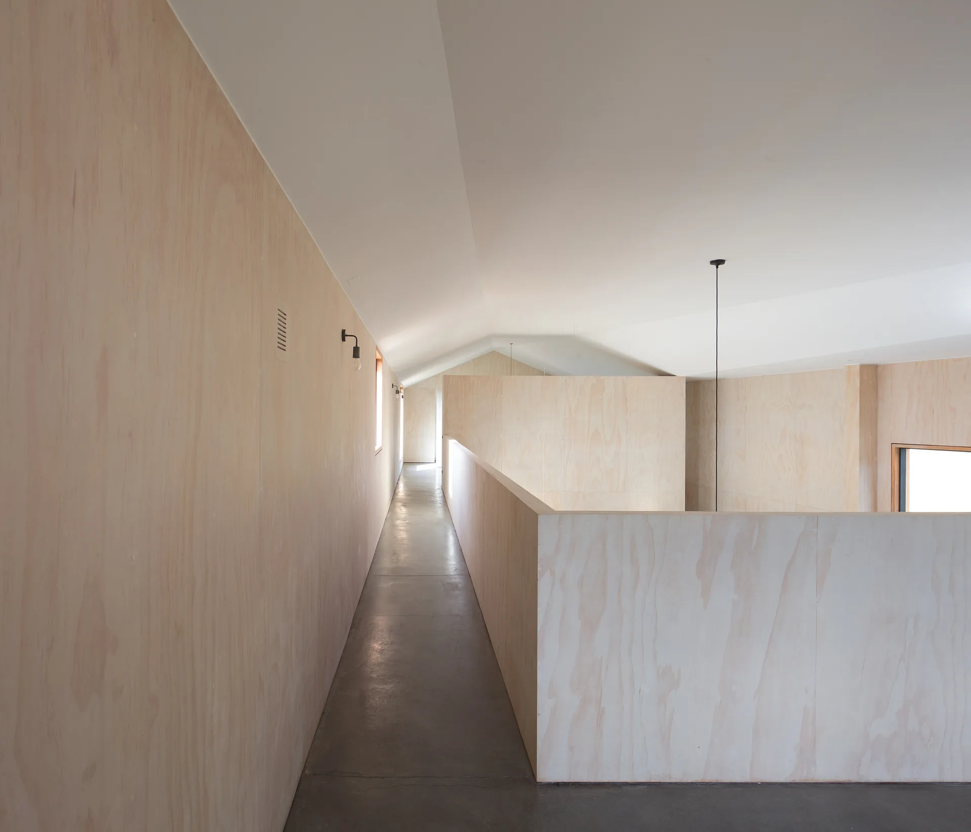 A minimalist residential hallway features light beige plywood wall paneling, a polished grey concrete floor, and a sloped white ceiling.