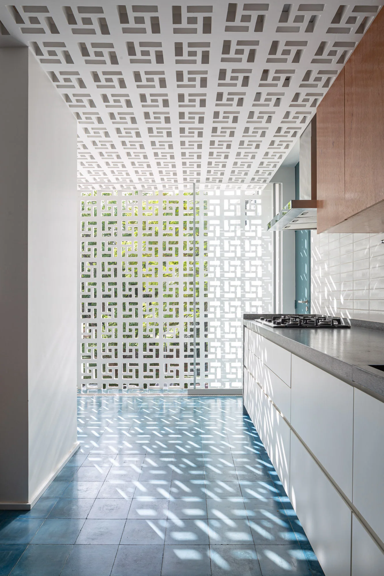 A modern kitchen features white base cabinets, wood upper cabinets, a blue tiled floor, and a white geometric breeze block screen casting sunlit patterns.