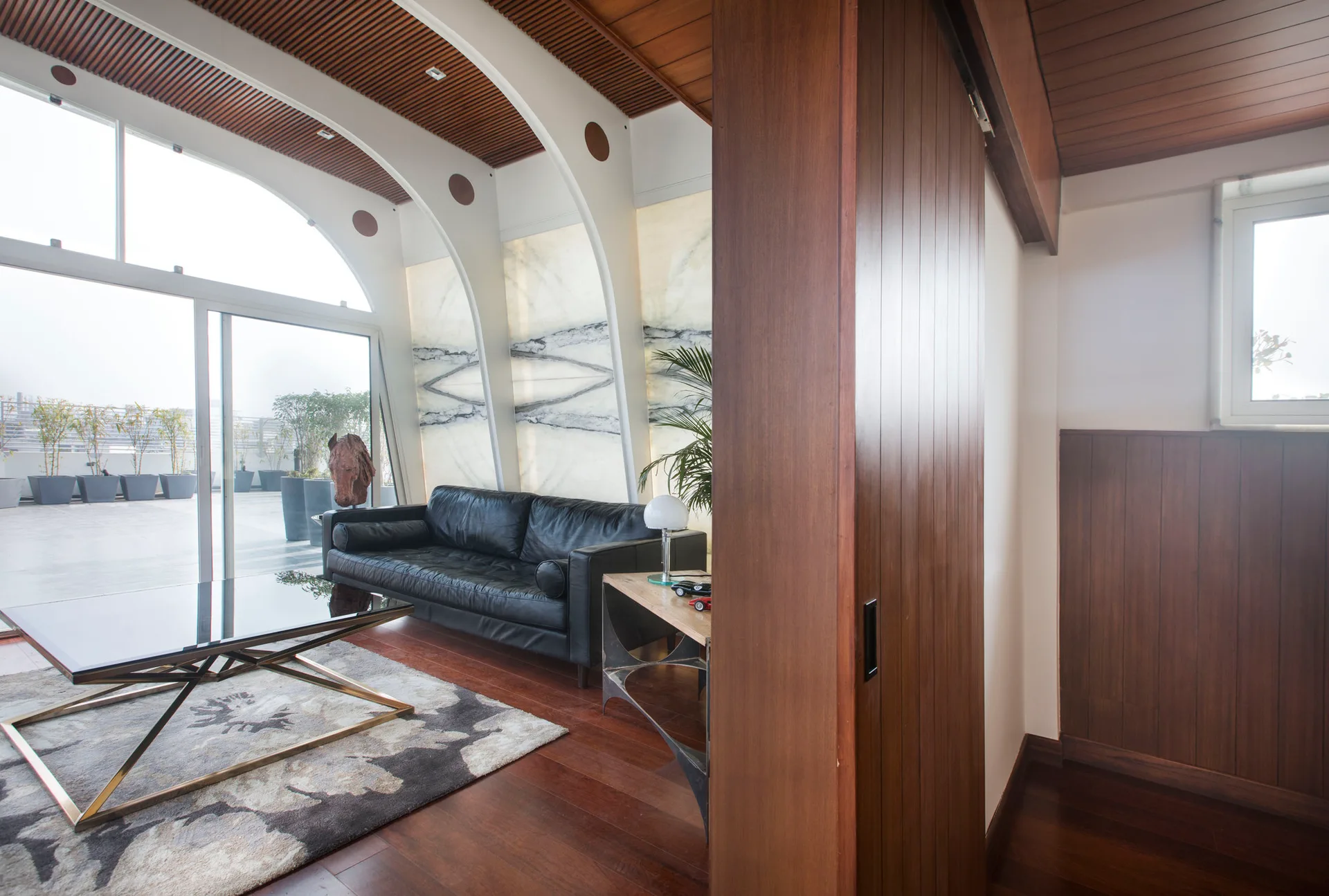 A living room featuring a black leather sofa, a geometric coffee table, and backlit marble wall panels under white arched ceiling beams.