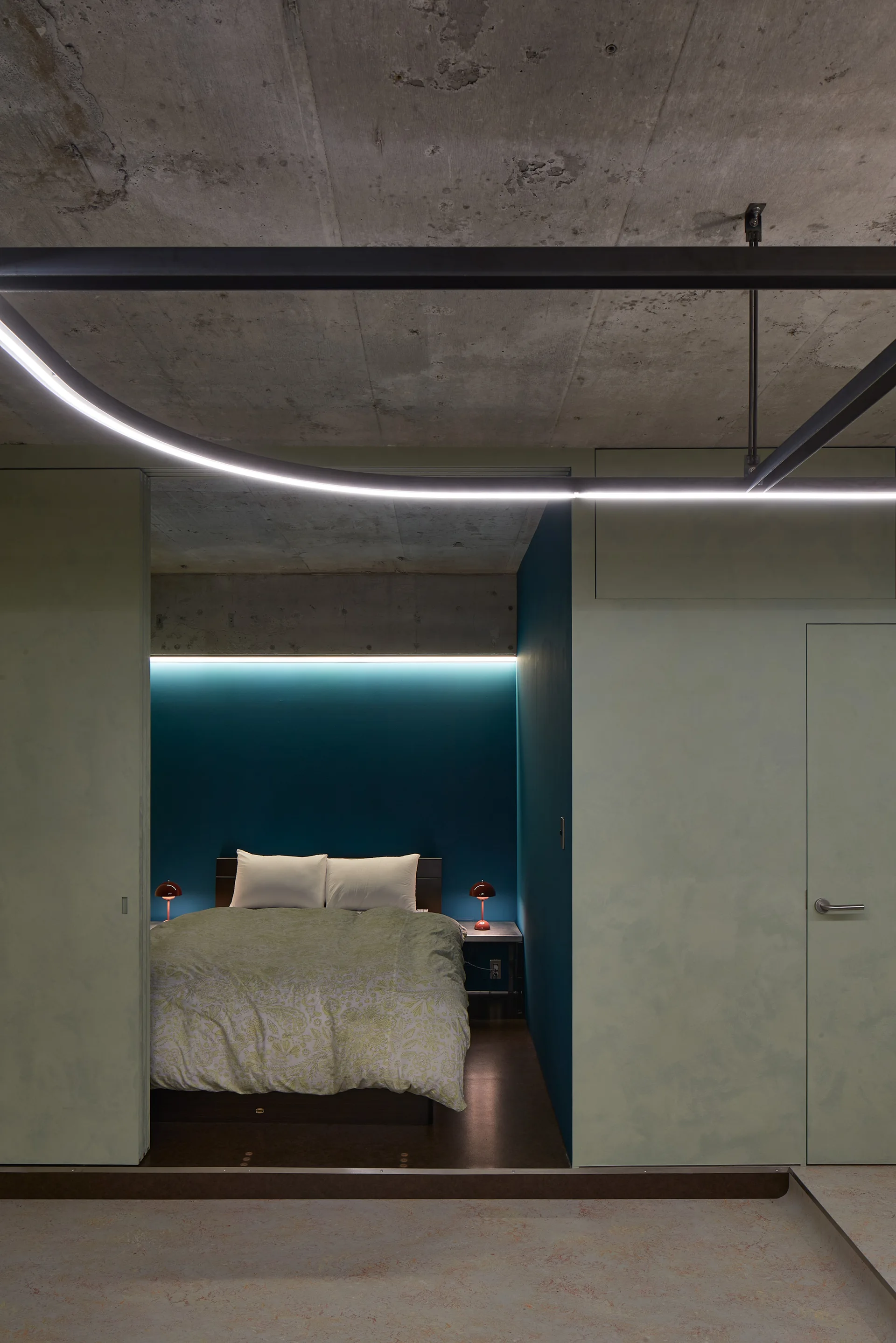 A modern bedroom features a bed with patterned linens against a deep teal wall beneath an exposed concrete ceiling and a curved ceiling light.
