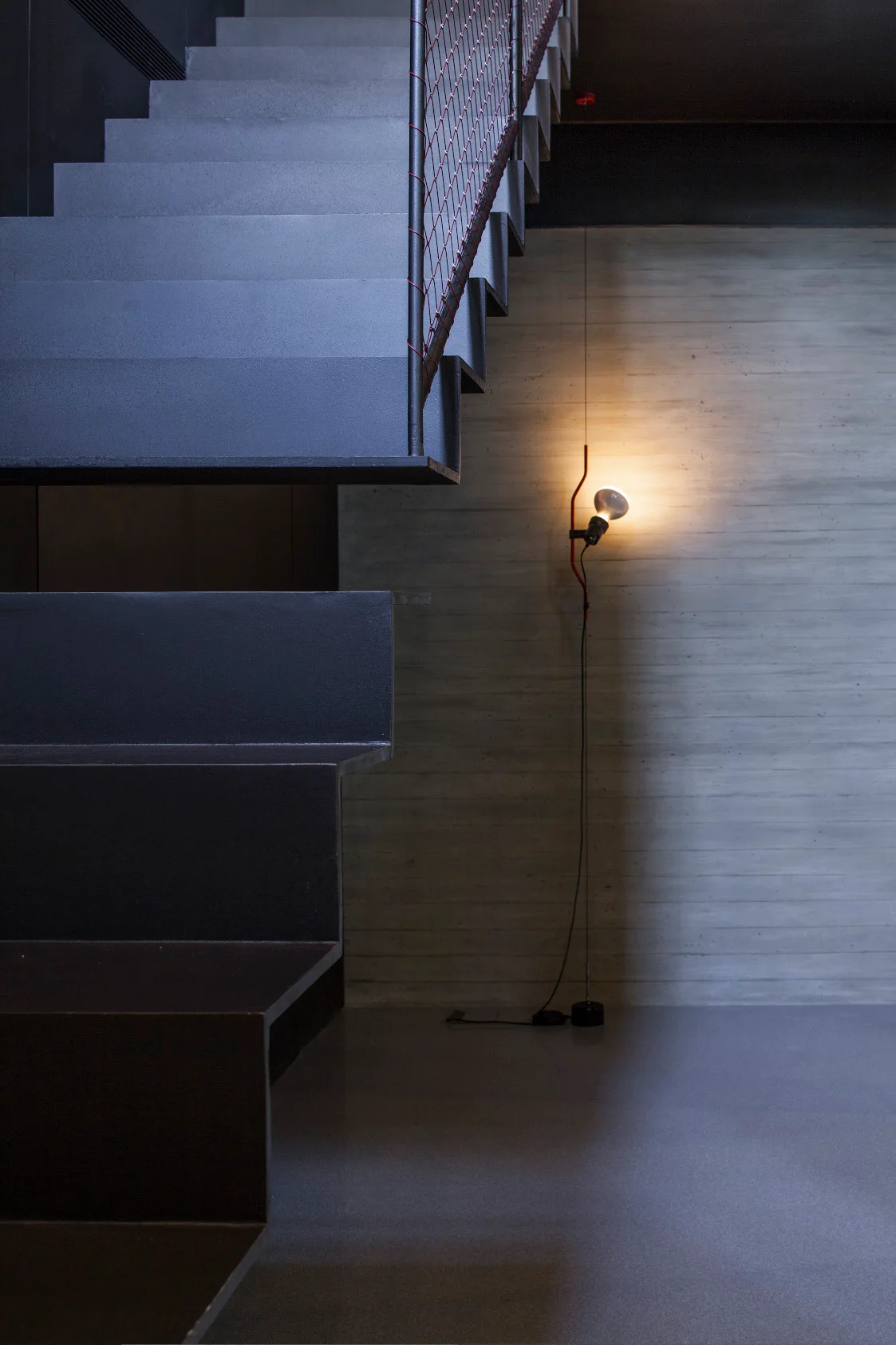 Dark metal stairs ascend past a textured concrete wall illuminated by a minimalist tension floor lamp.