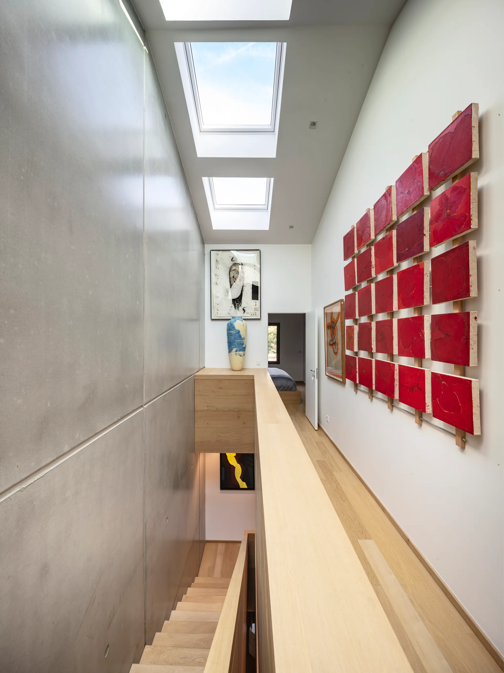 A bright modern hallway features a smooth concrete wall, a light wood staircase railing, dual ceiling skylights, and a grid of red wall art.