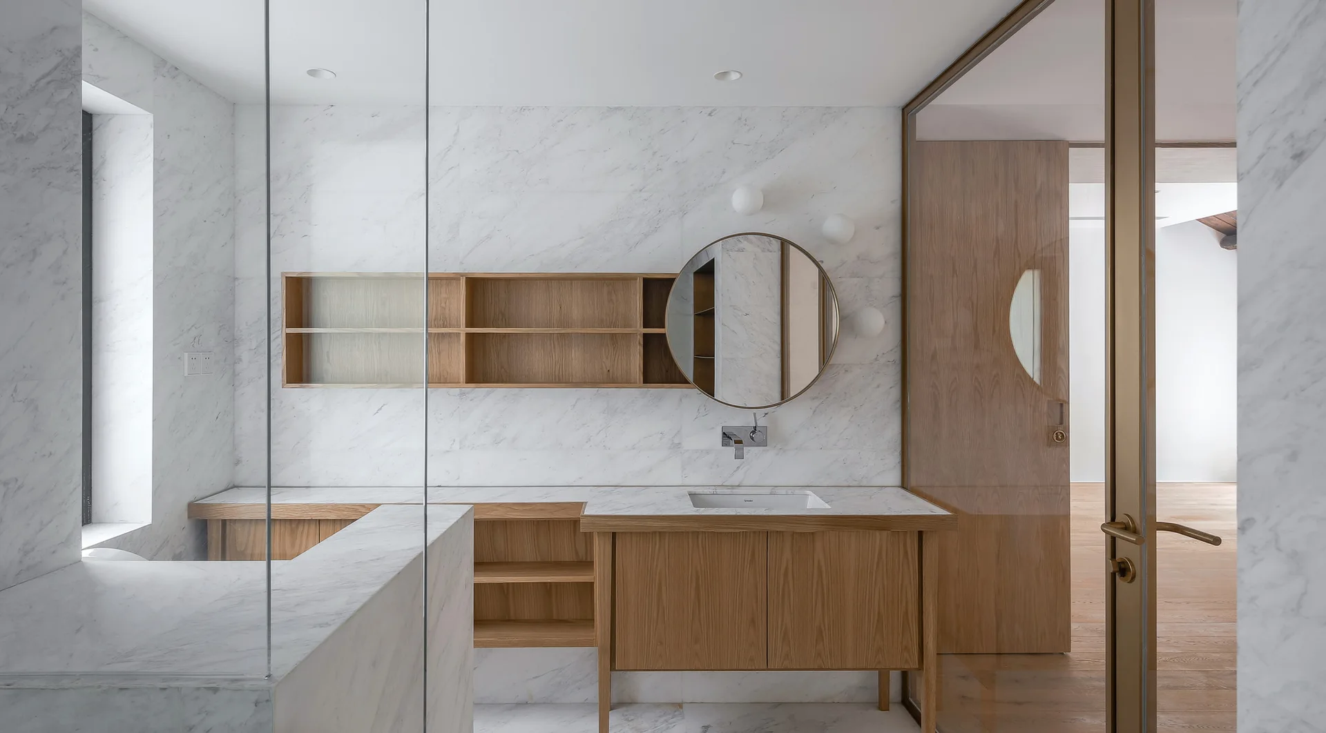 A contemporary bathroom features a wood and marble vanity, integrated open shelving, a large round mirror, and white veined marble walls.