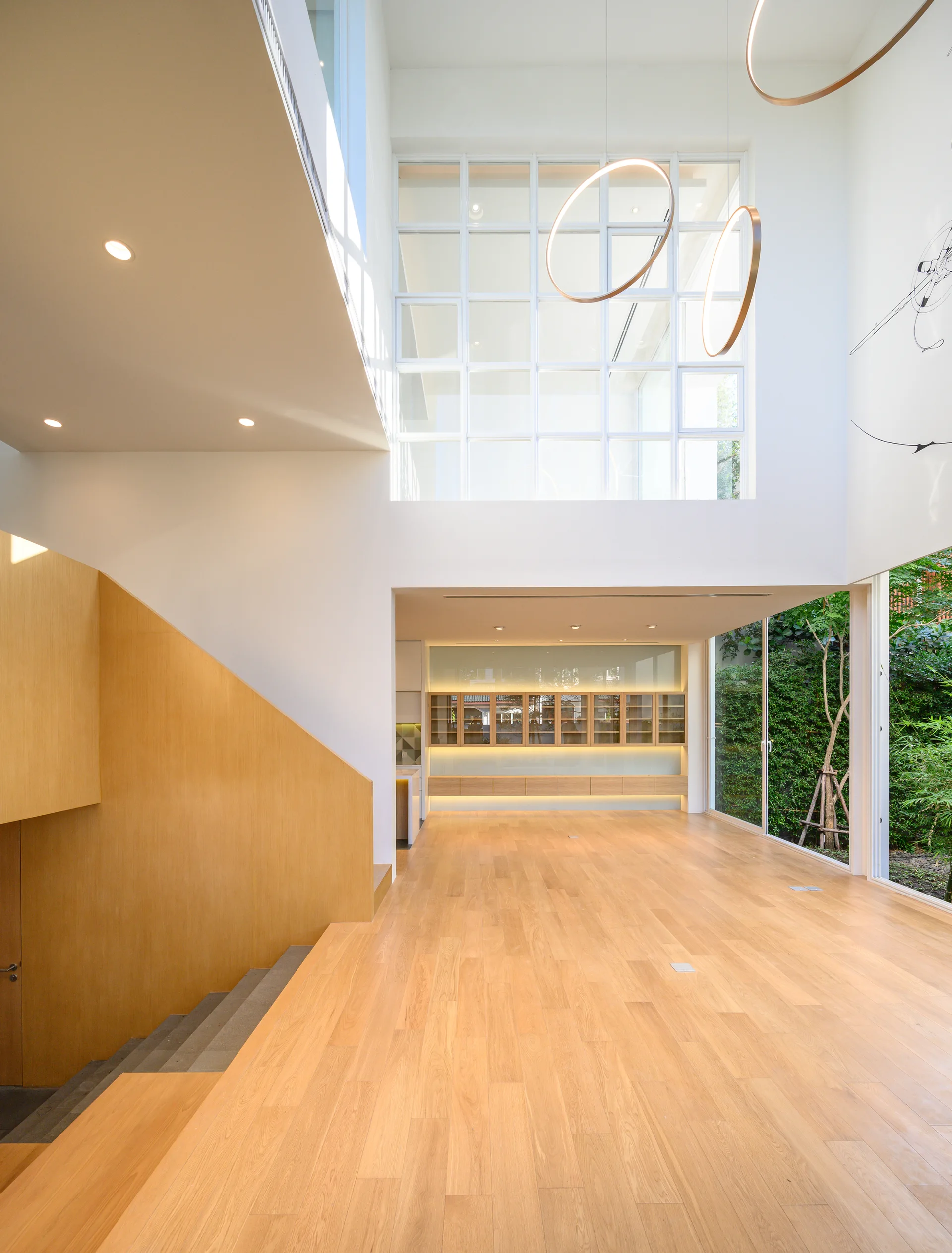 A double-height minimalist room features light wood flooring, a solid wood-paneled staircase, and a large grid window overlooking a garden.