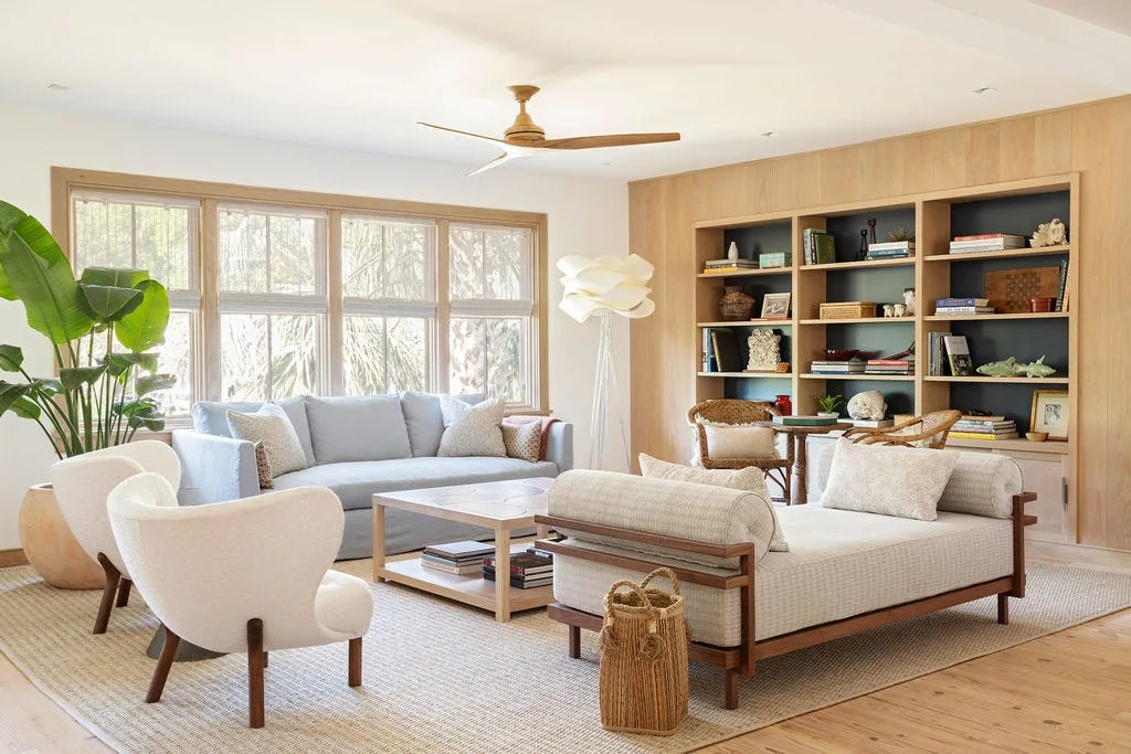 A bright living room features a light blue sofa under a window, white curved armchairs on a woven rug, and a large custom wood bookcase.
