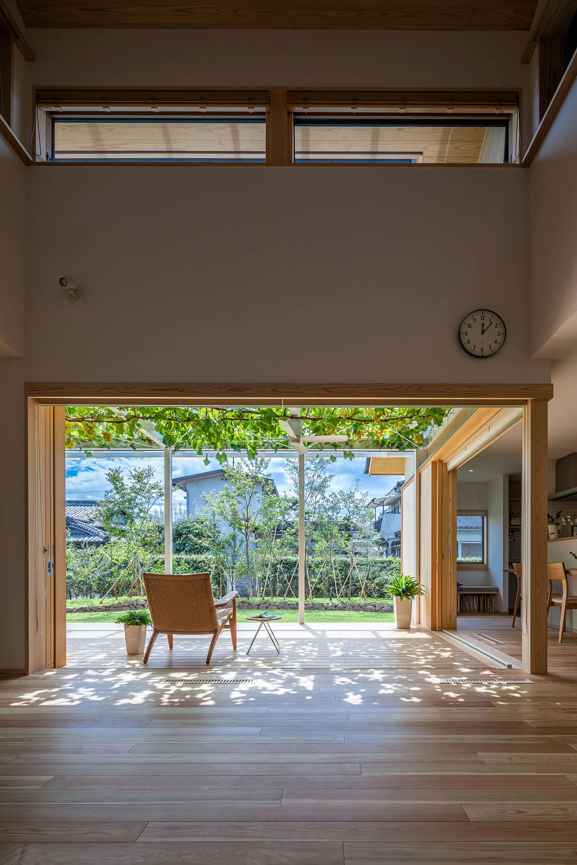 A minimalist living room features light wood floors and large open sliding glass doors leading to a sunlit garden terrace with a rattan armchair.