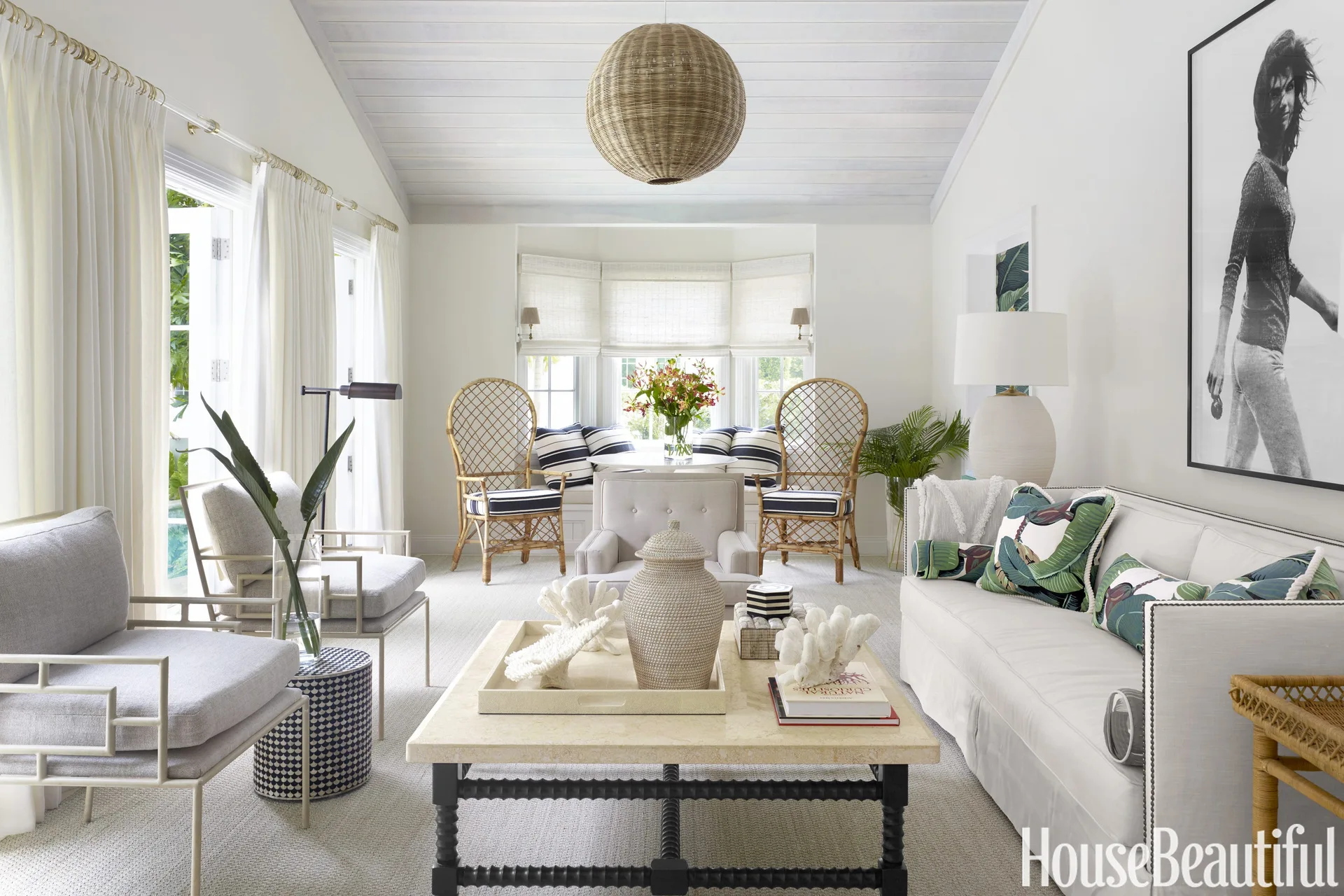 A bright coastal living room featuring a white sofa, a stone coffee table, tall rattan chairs, and a vaulted whitewashed wood ceiling.