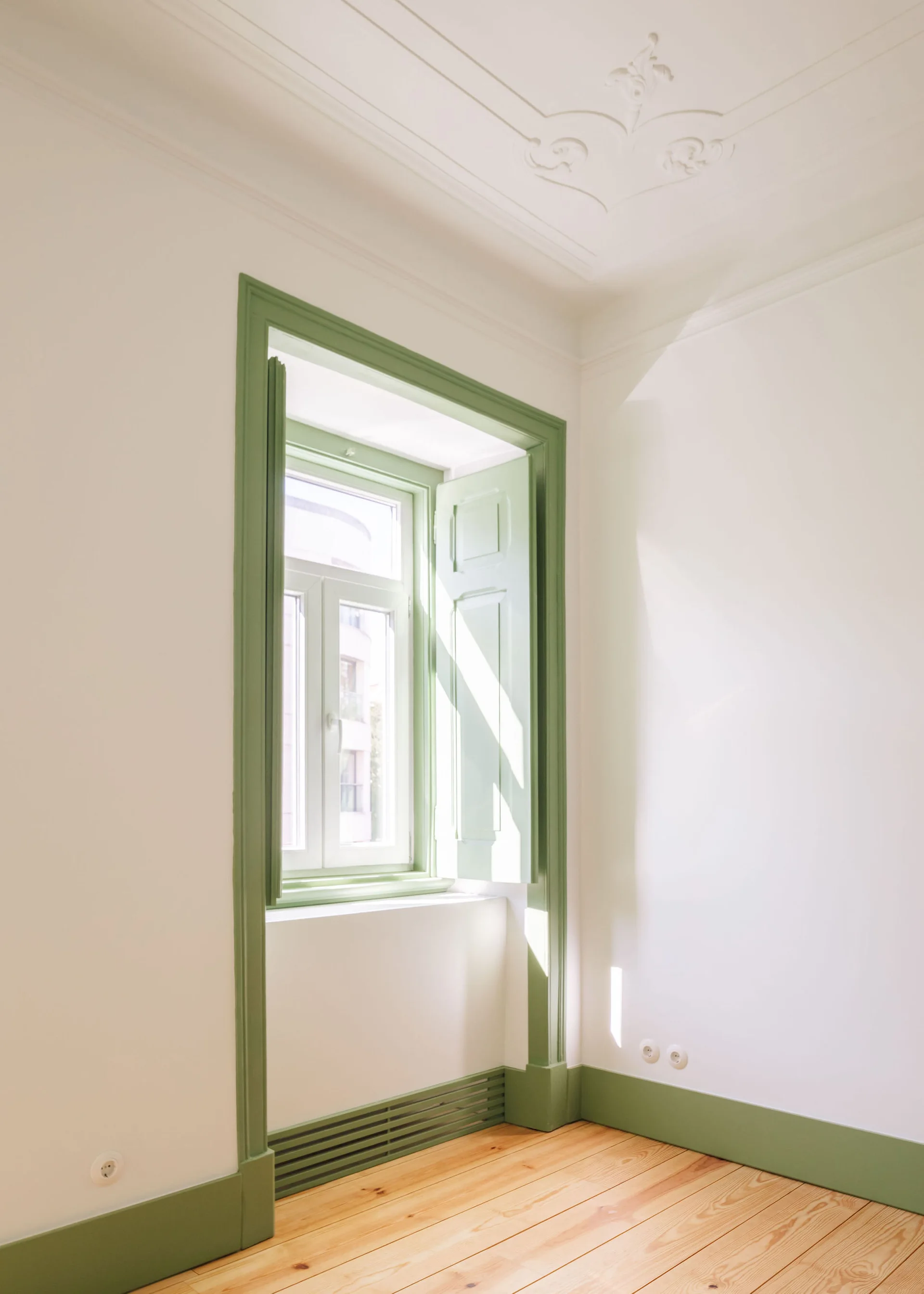 A bright room with white walls, light wood flooring, ornate ceiling molding, and a window with sage green painted shutters, trim, and baseboards.