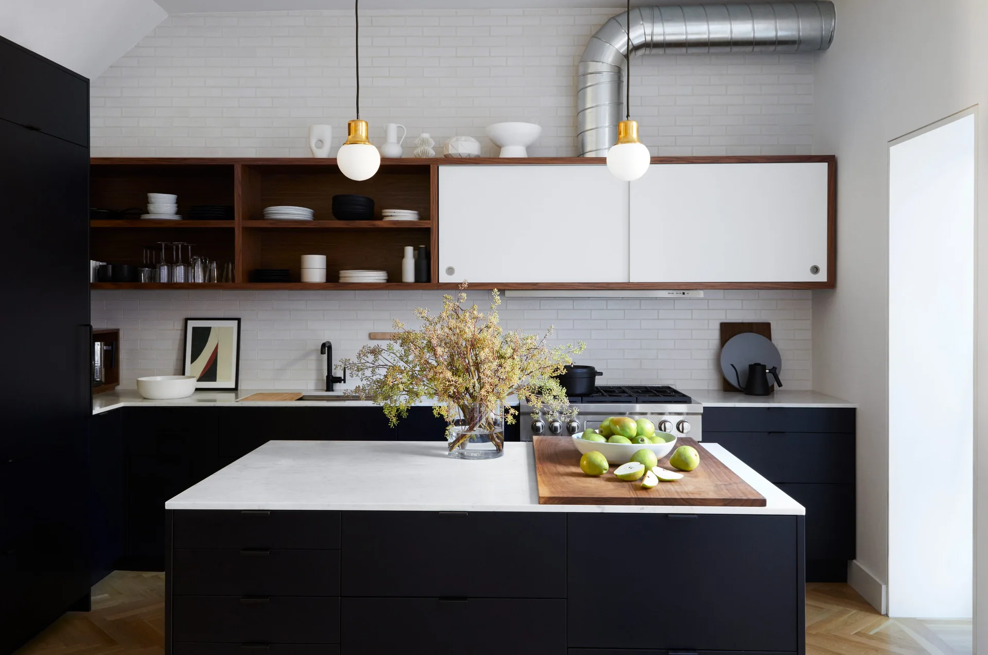 A modern kitchen features a black island with a white stone countertop, matte black lower cabinets, open wood shelves, and exposed metal ductwork.