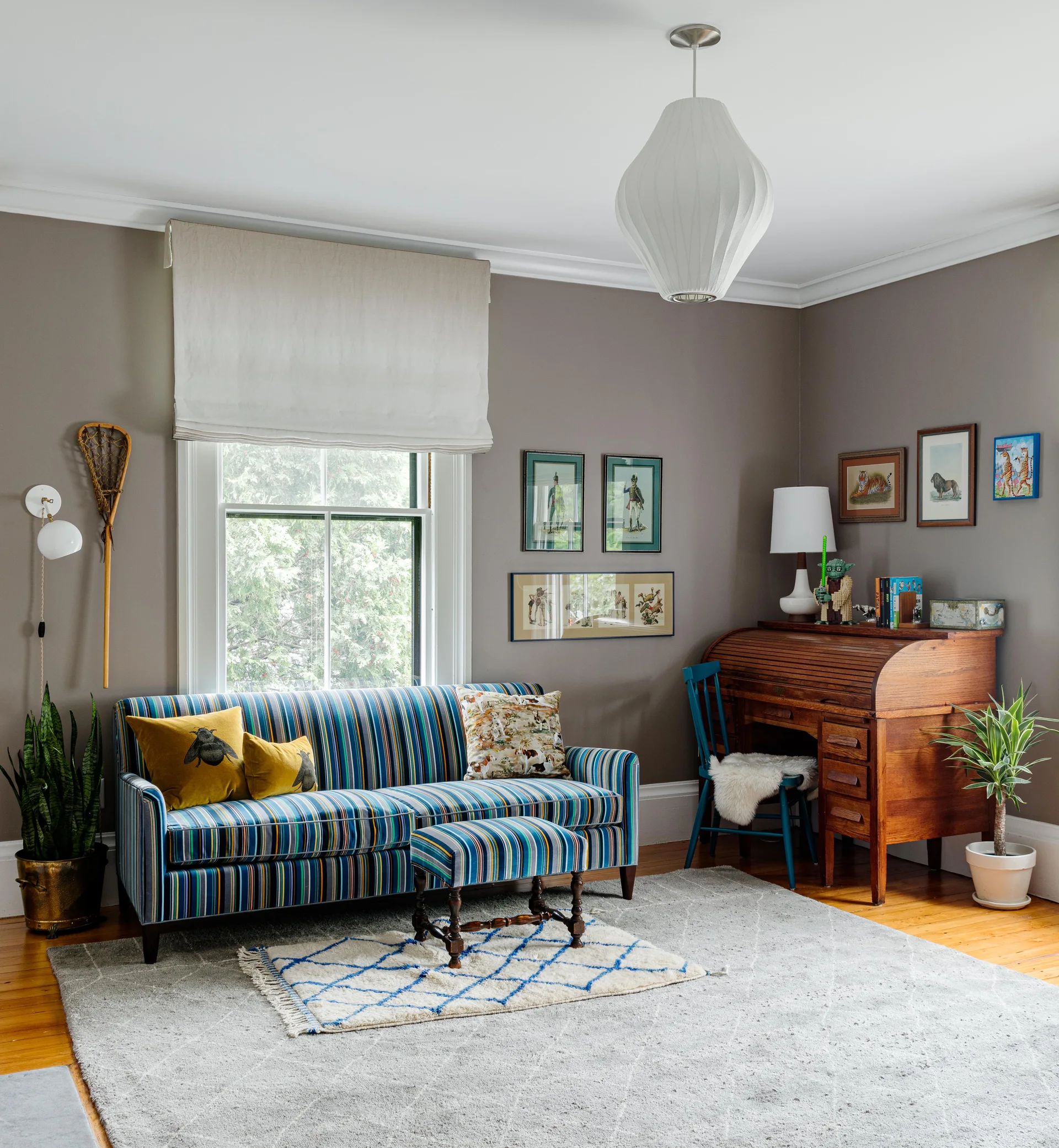 An eclectic living room features a blue striped sofa and matching ottoman near a vintage wooden roll-top desk on a grey area rug.
