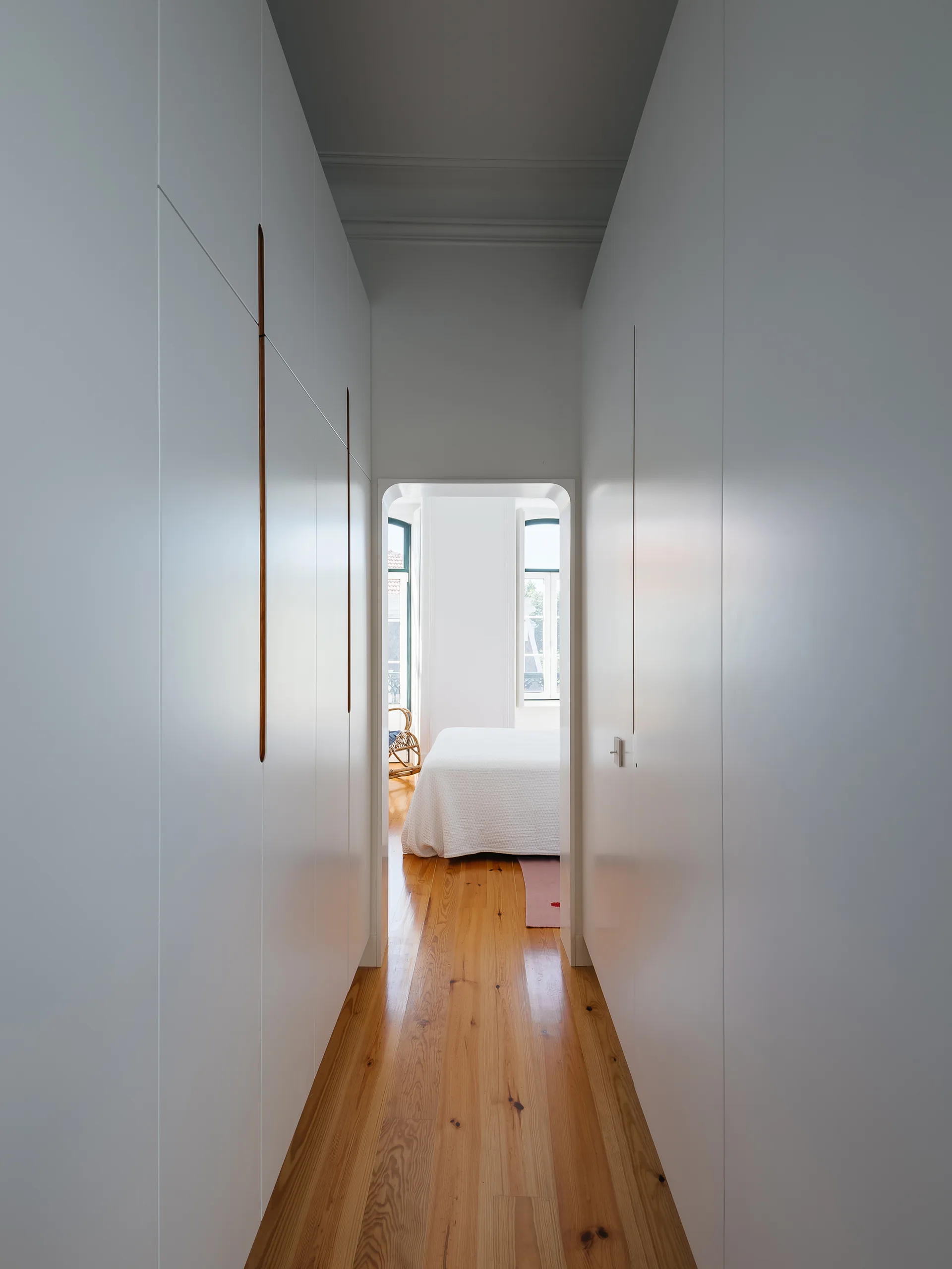 Built-in white floor-to-ceiling cabinets line a narrow hallway with natural wood floors leading to a sunlit bedroom through an arched doorway.