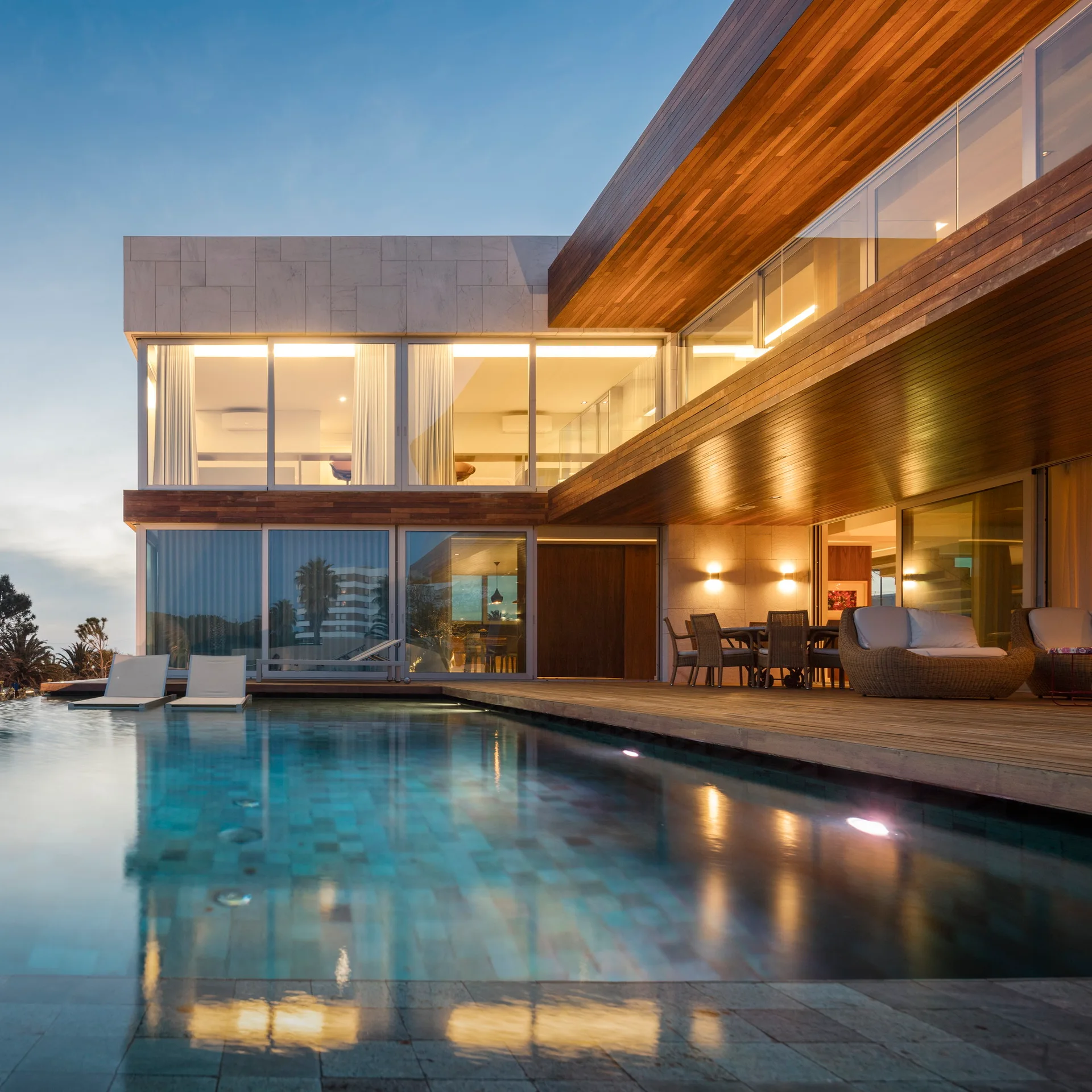 A modern two-story house features a wooden deck with outdoor furniture next to a swimming pool under a blue dusk sky.