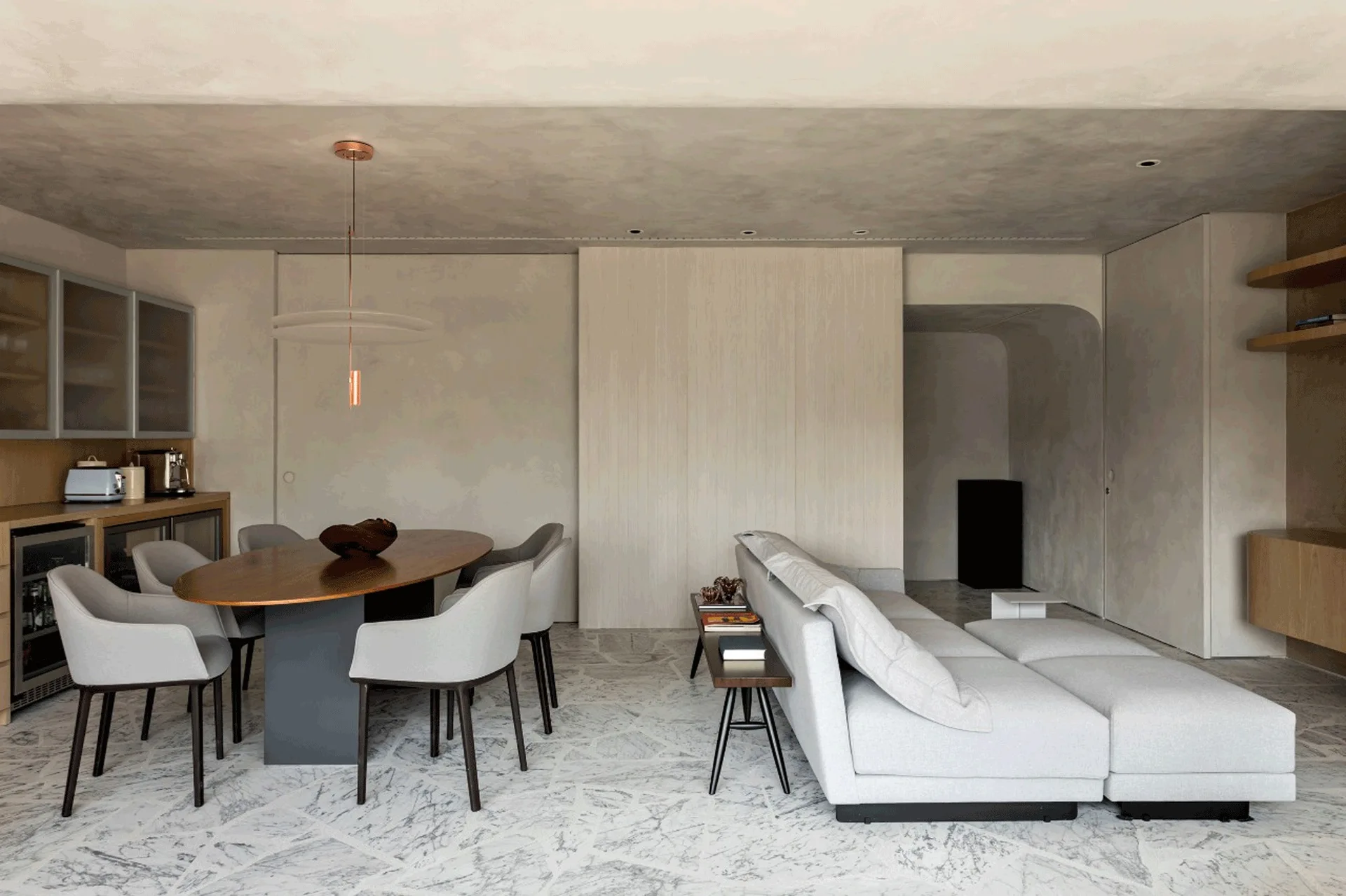 A minimalist open-concept living and dining space features a grey sectional sofa, an oval wooden table with chairs, and patterned white marble flooring.