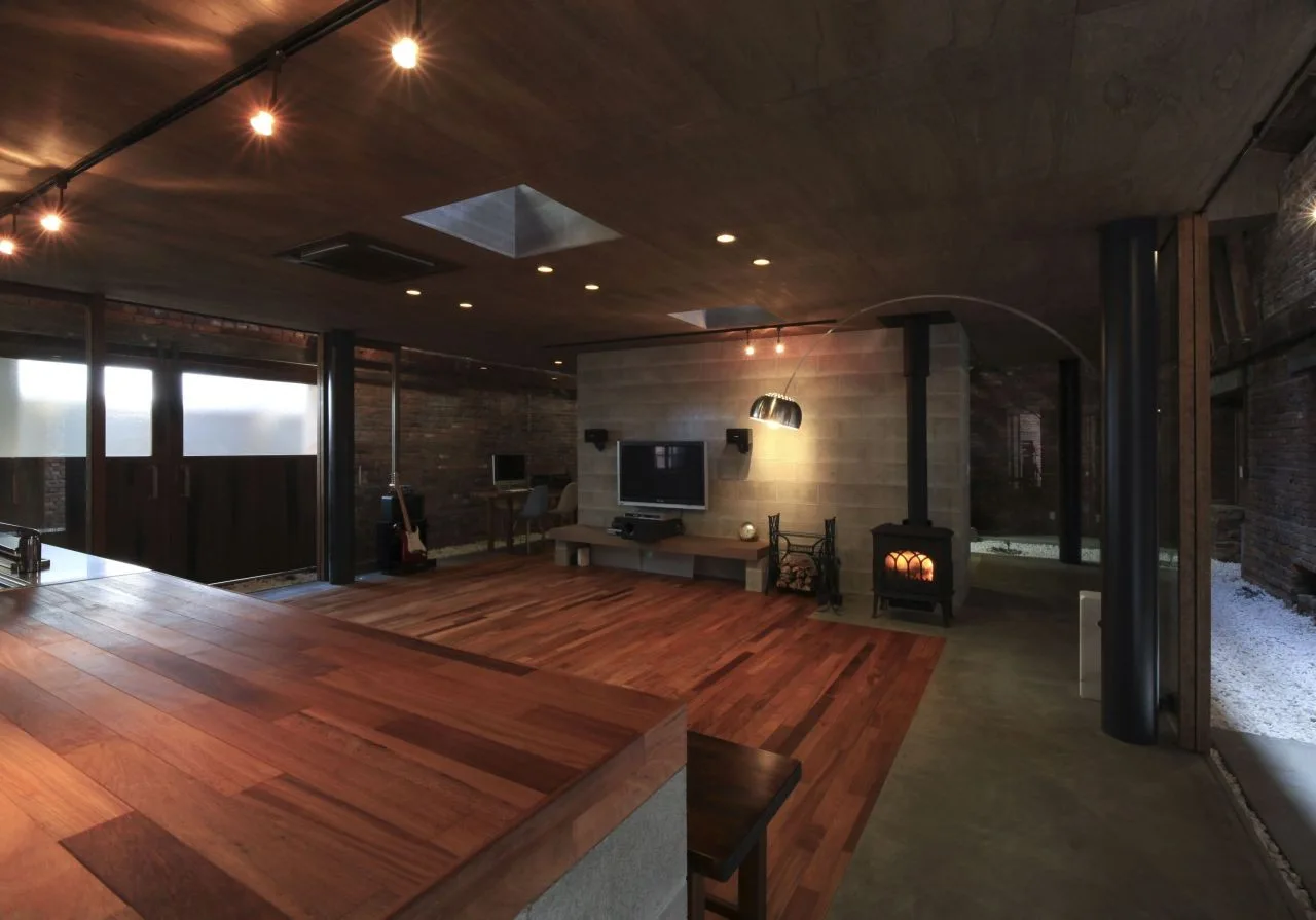 An industrial living room features a dark wood paneled ceiling, hardwood floors, a concrete block media wall, and a black wood-burning stove.