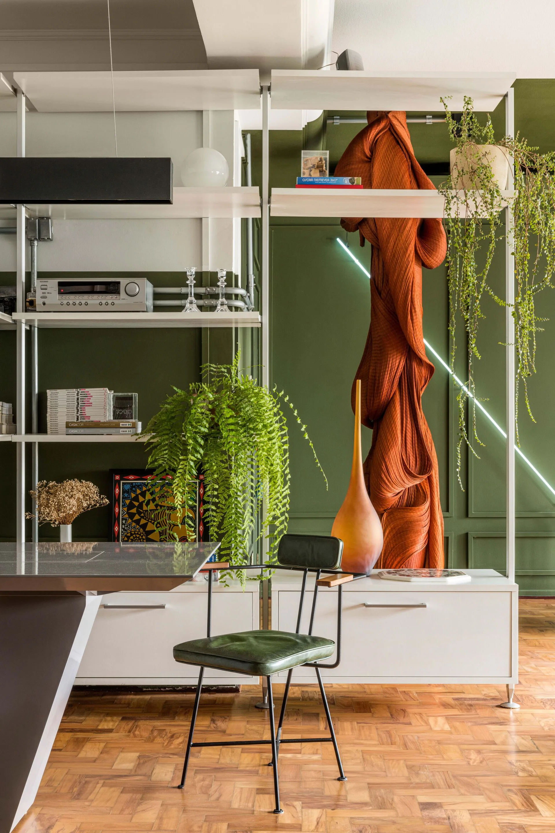 A green leather chair sits on parquet flooring in front of white modular shelving and an olive green paneled wall with a diagonal light.