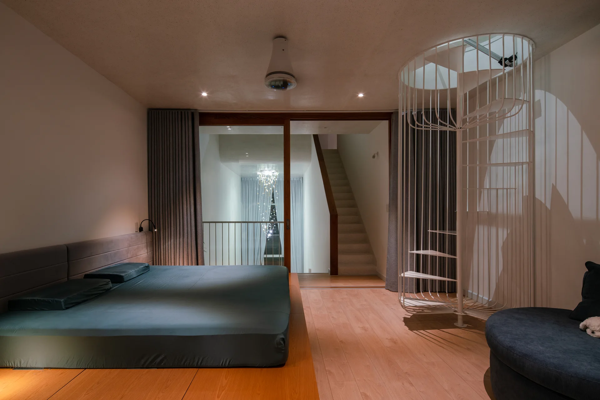 A modern bedroom features a low platform bed with grey sheets, wood flooring, a white metal spiral staircase, and glass doors opening to a hallway.