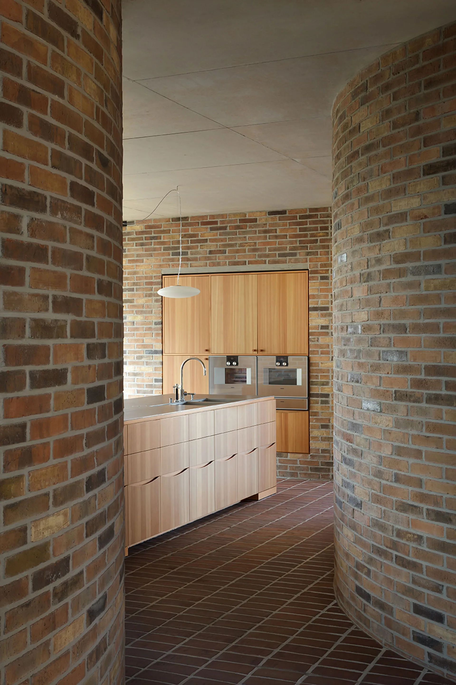 A view between curved brick walls looking into a modern kitchen with a wooden island, built-in cabinetry, and integrated appliances.