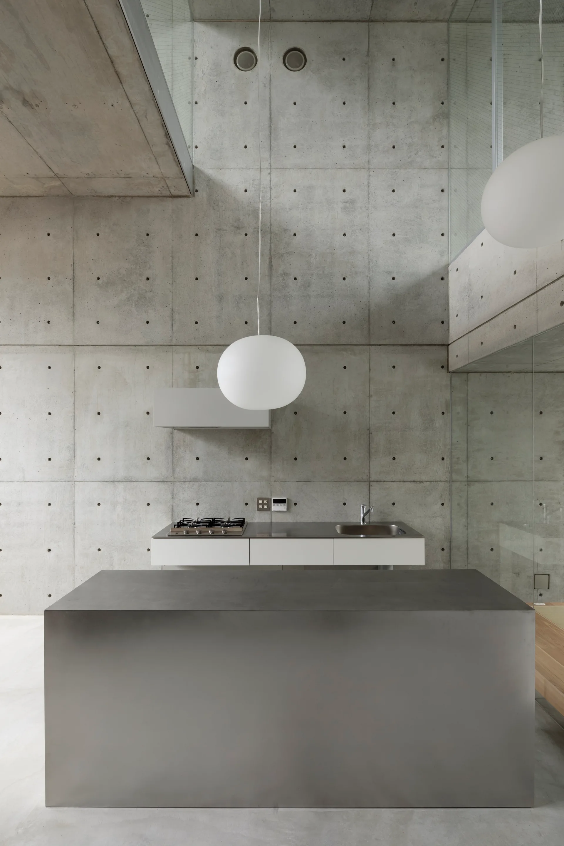 A minimalist kitchen features a large grey metal island, a white counter with a sink and stove, exposed concrete walls, and a round white pendant light.