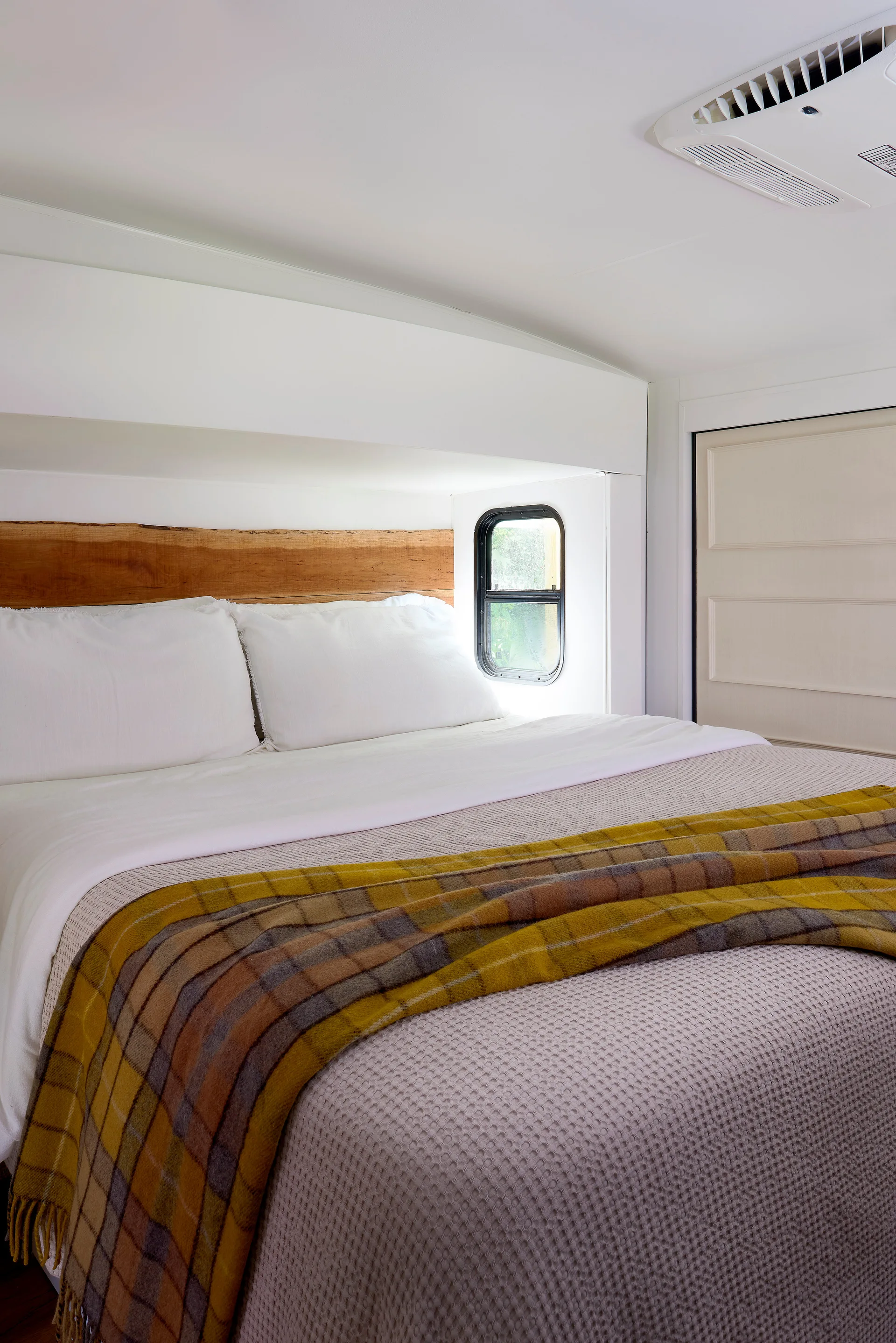 A compact bed in a white camper bedroom features a live-edge wood headboard, a textured grey bedspread, and a yellow plaid throw blanket.
