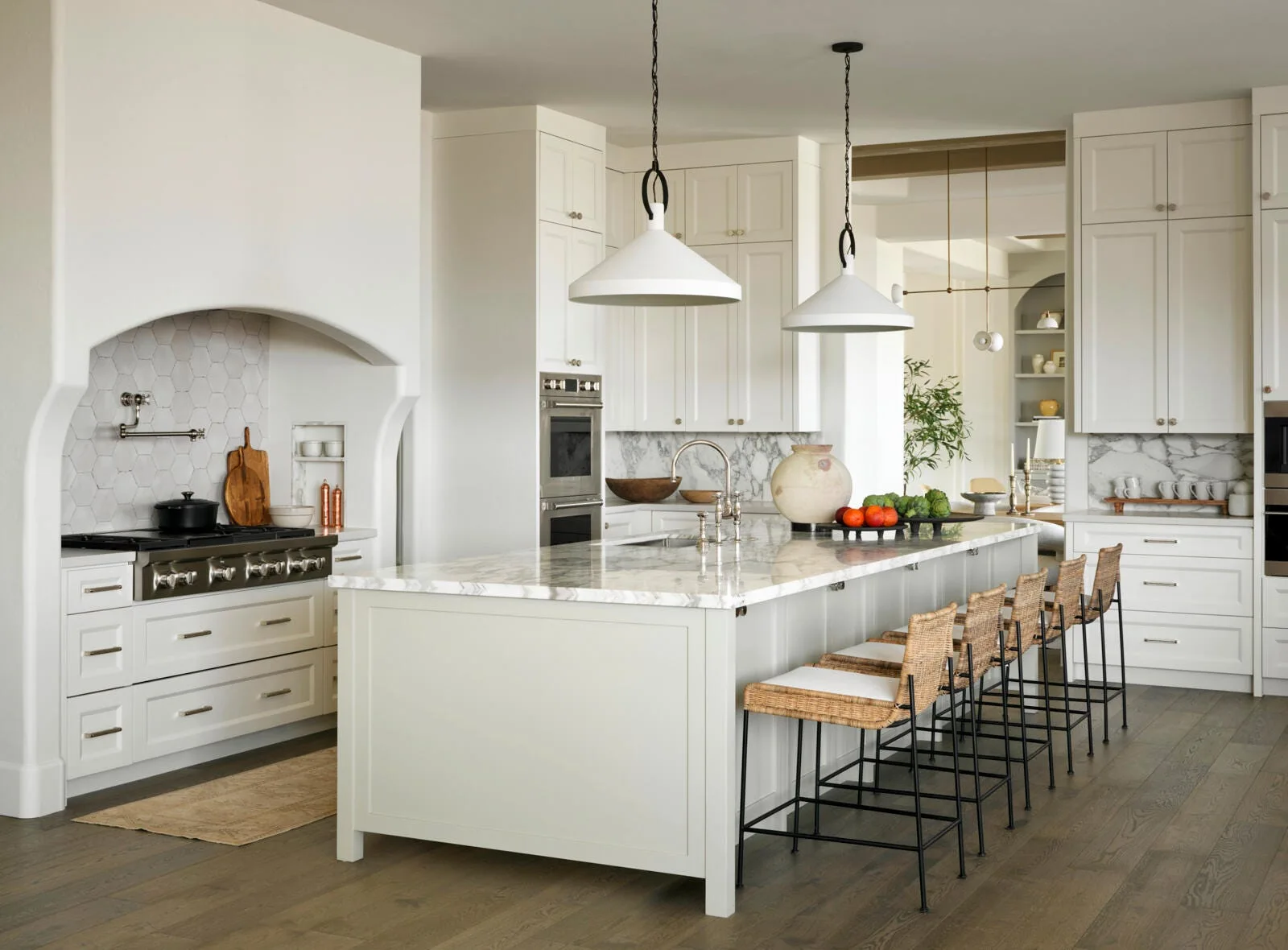 A bright kitchen features a marble-topped island with woven bar stools, white cabinetry, and an arched alcove built around a stainless steel range.