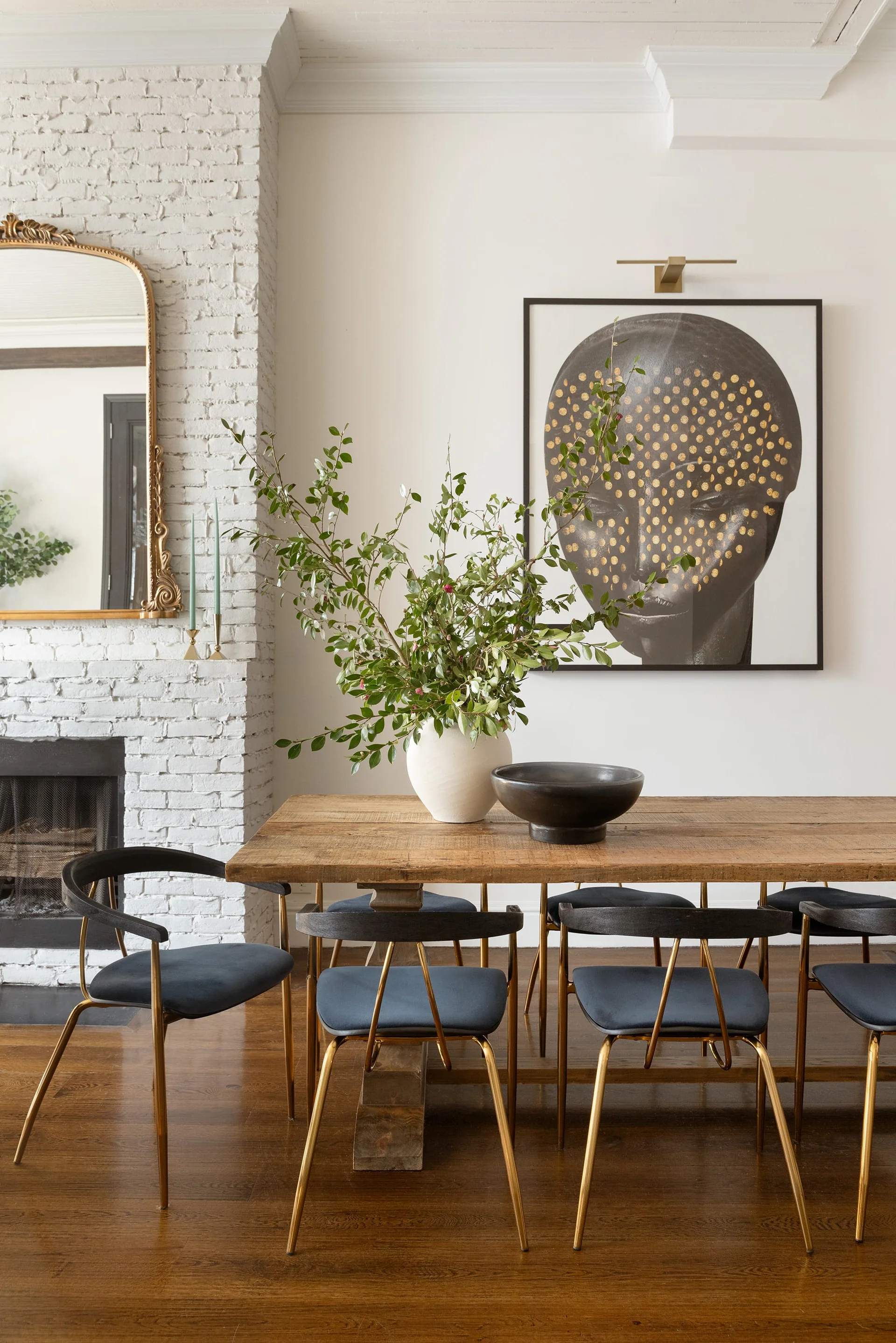 A rustic wooden dining table surrounded by black chairs with brass legs sits near a white painted brick fireplace and large black wall art.