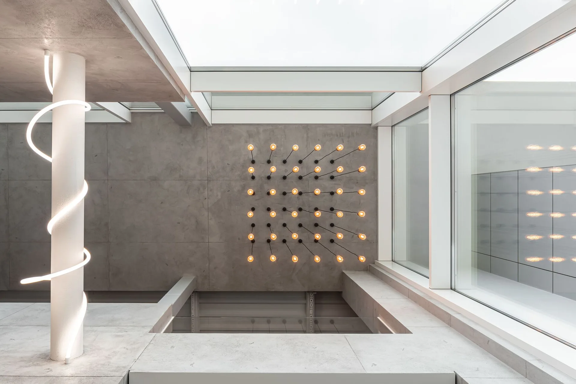 A minimalist atrium features large-format grey concrete panels on the walls, a spacious glass skylight, and a grid of exposed warm-glowing light bulbs.