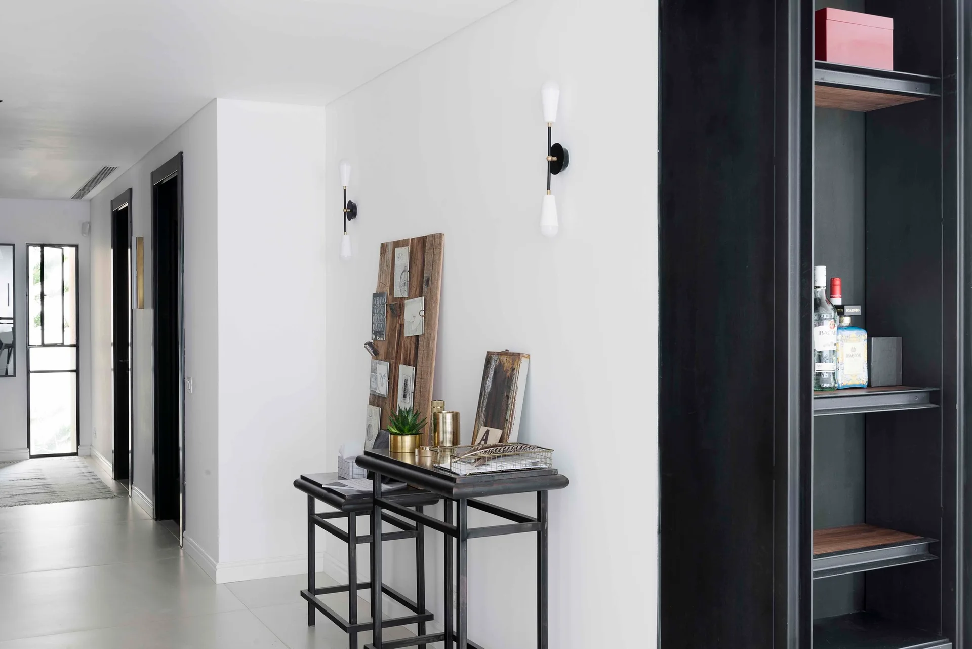 A bright white hallway with a black metal console table, modern wall sconces, and a tall black built-in shelving unit.