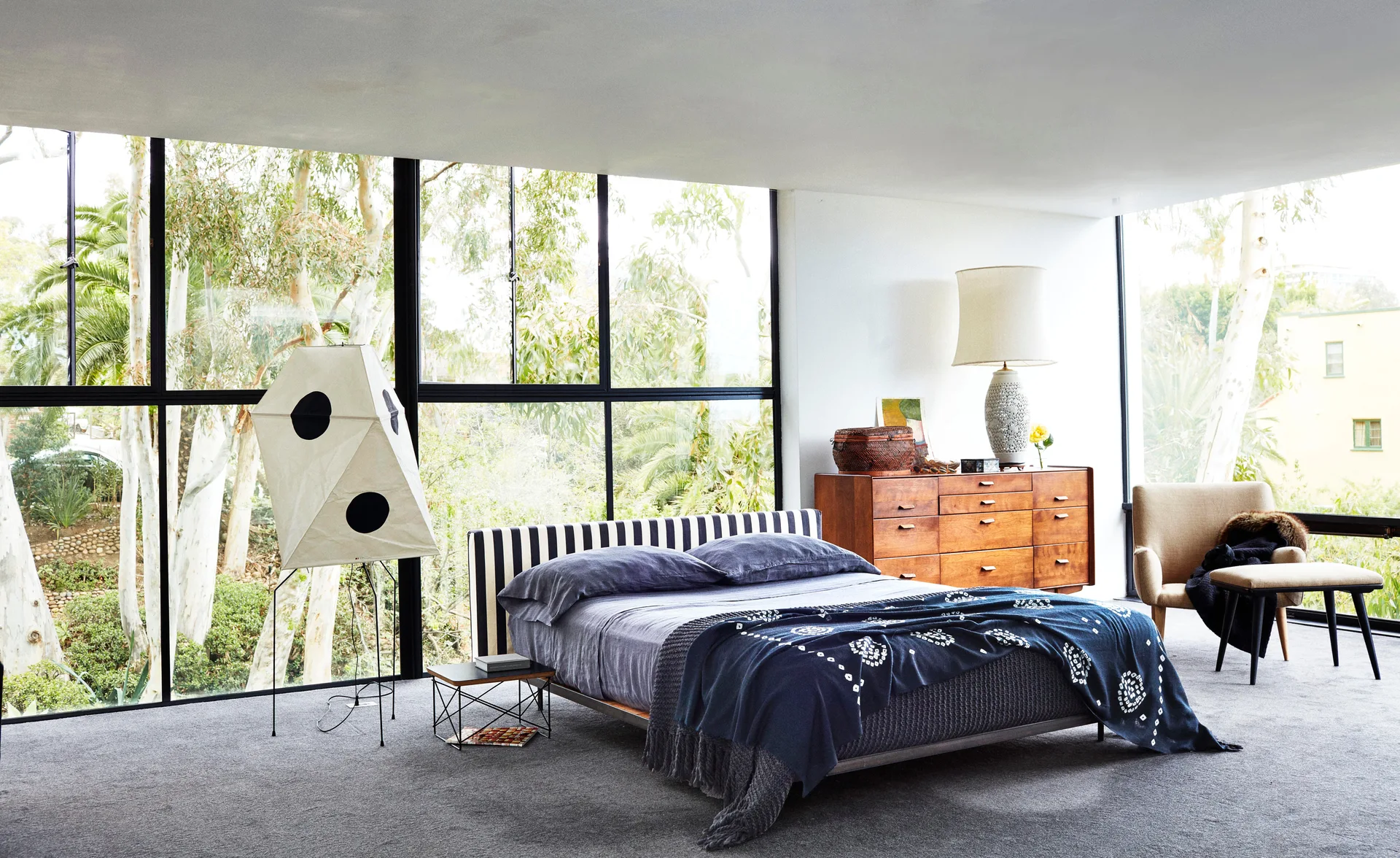 A modern bedroom features a bed with a striped headboard, a wooden dresser, and floor-to-ceiling windows looking out at green trees.
