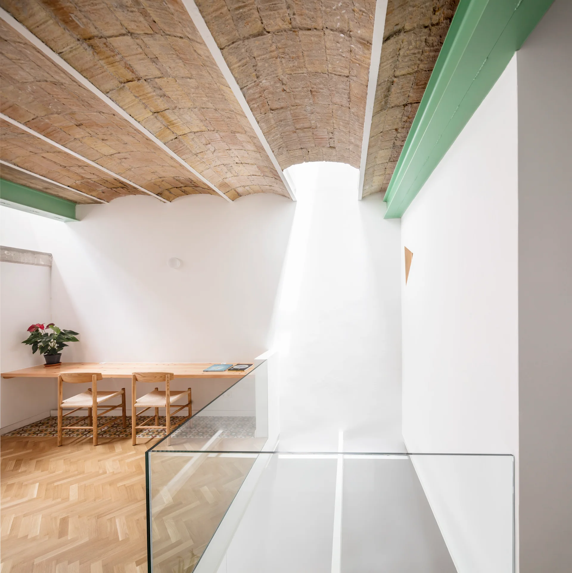 A bright loft workspace features a long wooden desk with two chairs under a vaulted brick ceiling next to a glass railing.