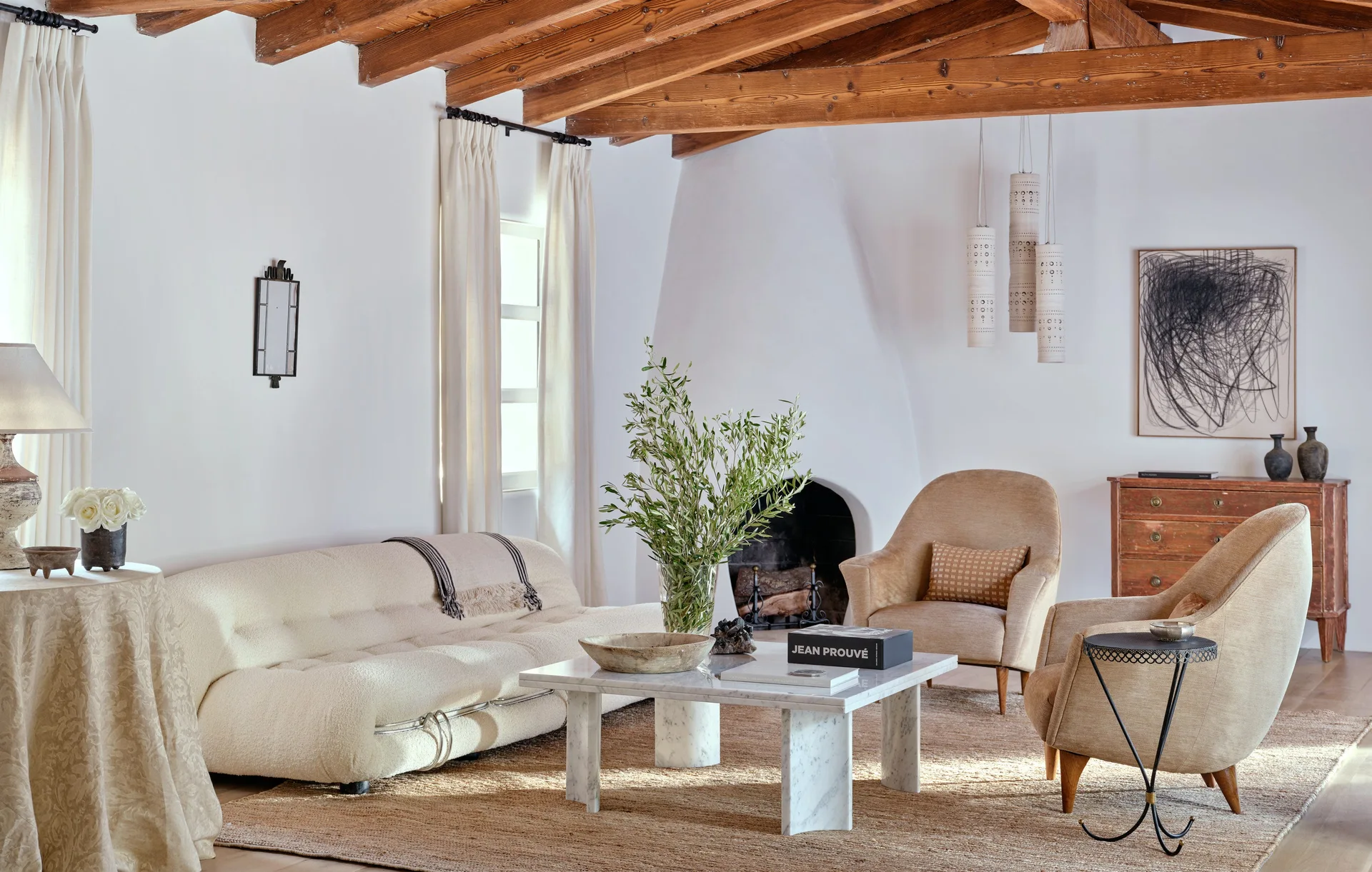 A bright living room features a textured cream sofa, a marble coffee table, and two tan armchairs near an arched plaster fireplace under exposed wood beams.