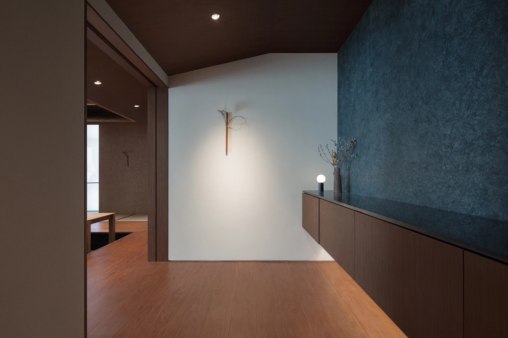 A minimalist hallway featuring a long wall-mounted wooden cabinet, wood floors, and a dark textured charcoal wall with under-cabinet lighting.