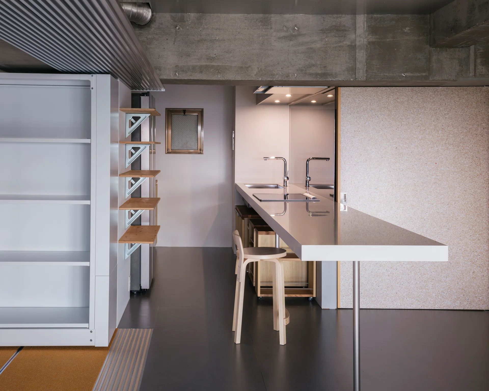 A minimalist kitchen features a long glossy white countertop with a wooden bar stool under an exposed concrete ceiling.