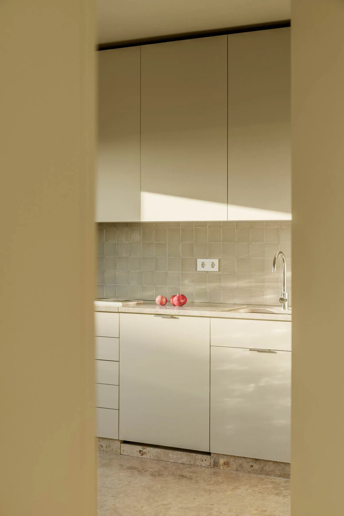 Minimalist beige kitchen featuring flat-front cabinets, a square-tile backsplash, stone countertops, and a chrome faucet viewed through a doorway.