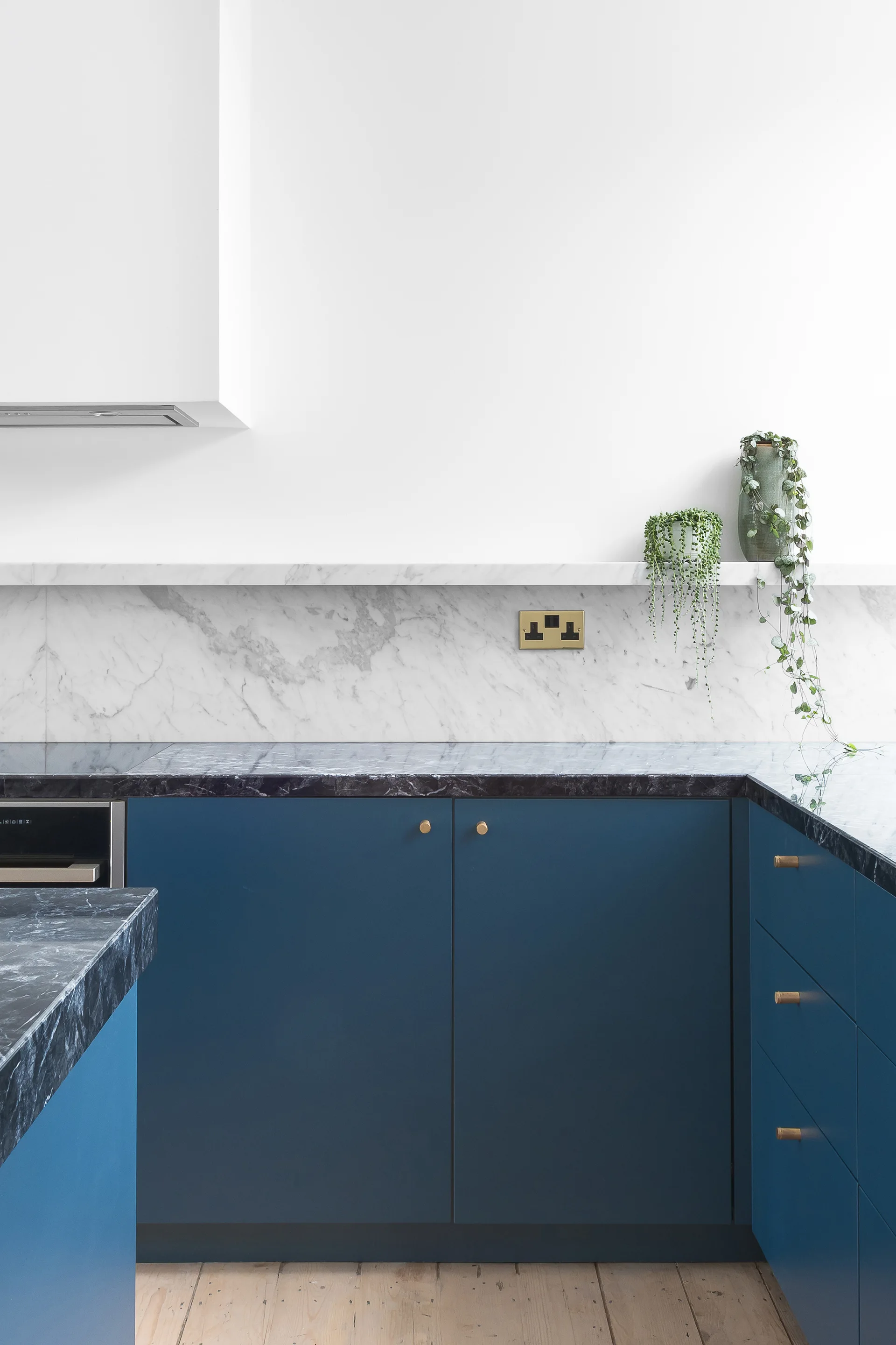 Modern kitchen corner showing matte blue cabinets with brass pulls, a dark veined marble countertop, a white marble backsplash, and a floating shelf.