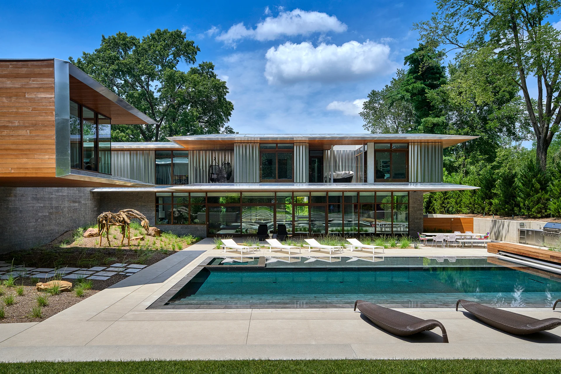 A modern two-story home with wood and metal siding faces a rectangular swimming pool surrounded by a concrete patio with lounge chairs.