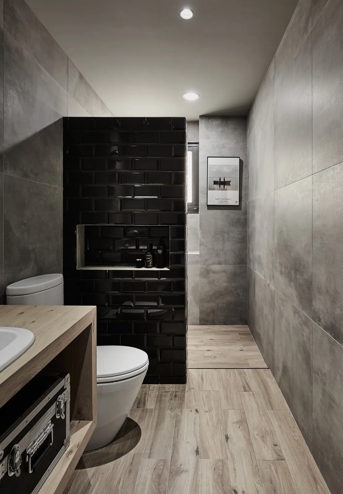 A modern bathroom featuring a glossy black subway tile partition wall, large grey concrete-look wall tiles, and light wood plank flooring.