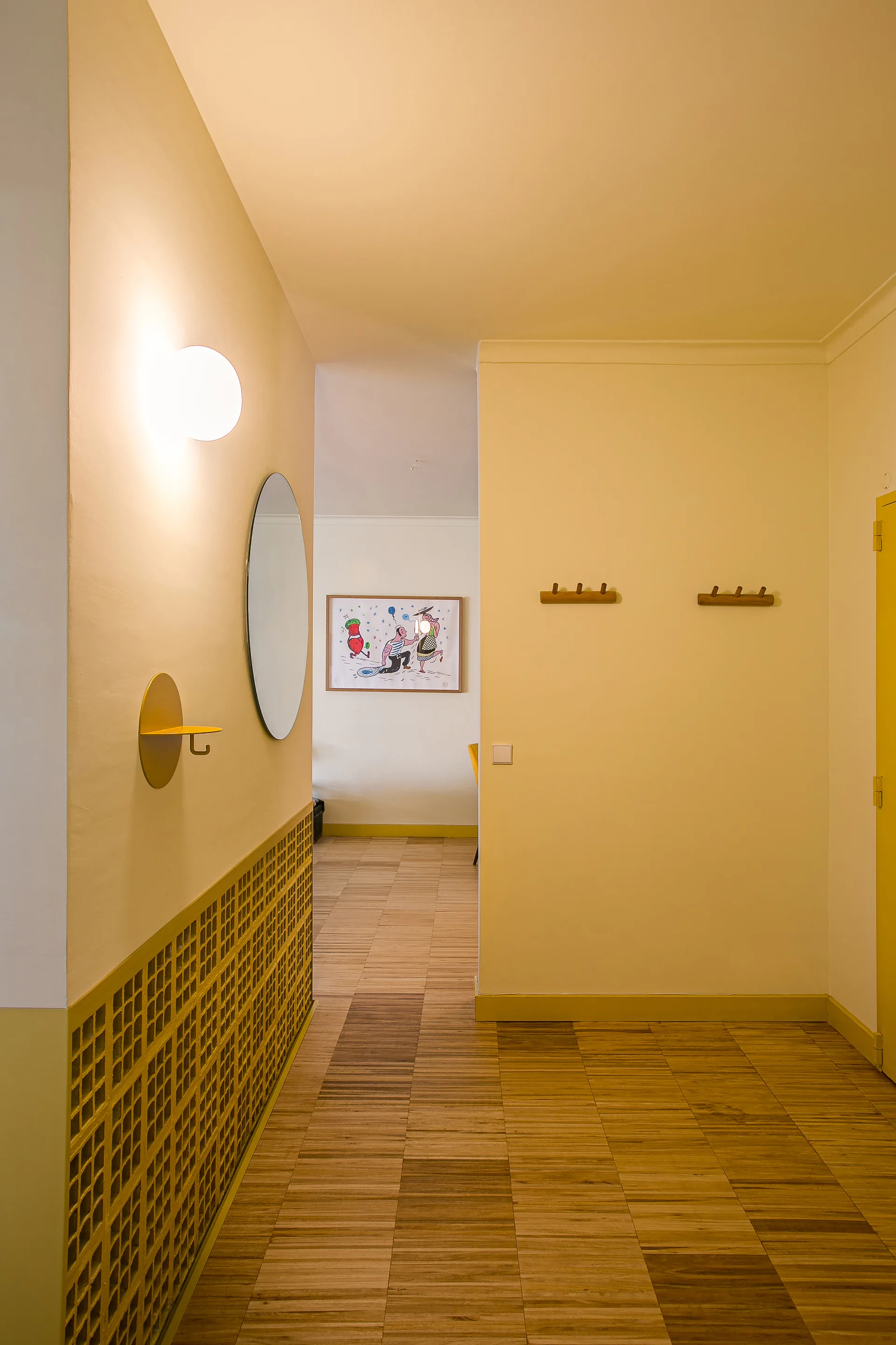 A bright yellow hallway features textured wood flooring, a circular wall mirror, a glowing globe sconce, and matching yellow grid wainscoting.