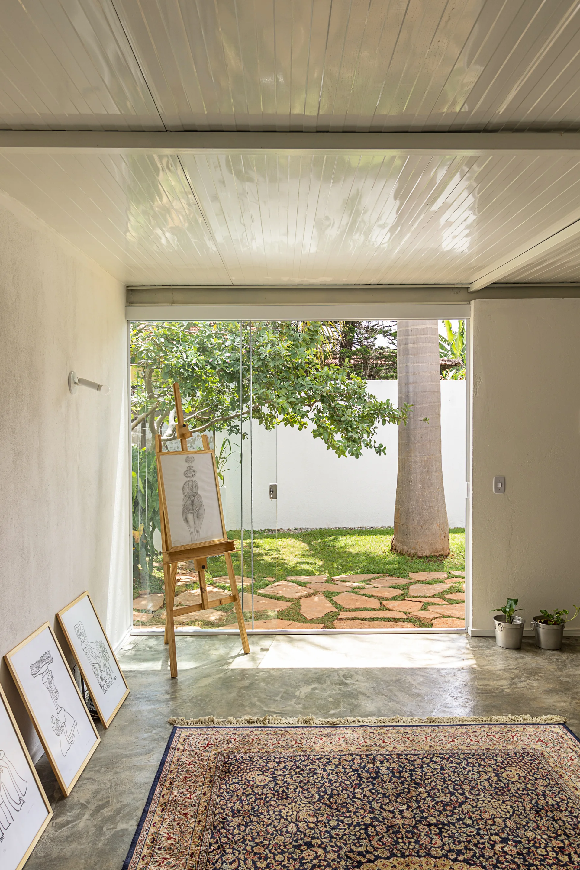 An art studio features a wooden easel, framed drawings leaning on a polished concrete floor, and a sliding glass wall opening to a green garden.