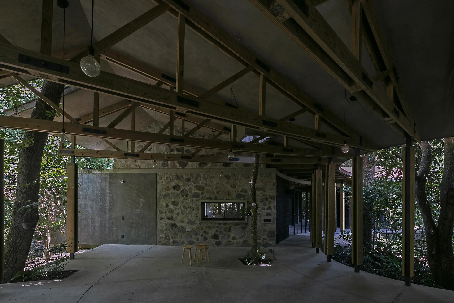 A covered outdoor patio features an exposed timber truss ceiling, stone and concrete walls, and living trees growing directly through the concrete floor.