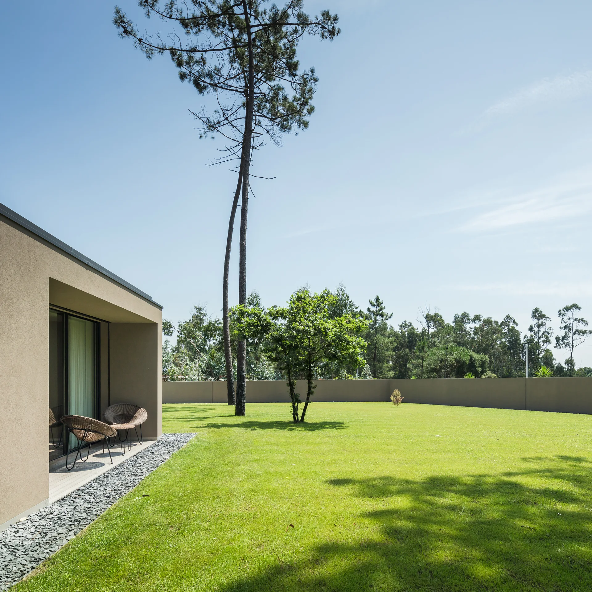 A minimalist beige house features a recessed patio with wicker chairs overlooking a vast green lawn bordered by a grey gravel bed and tall pine trees.