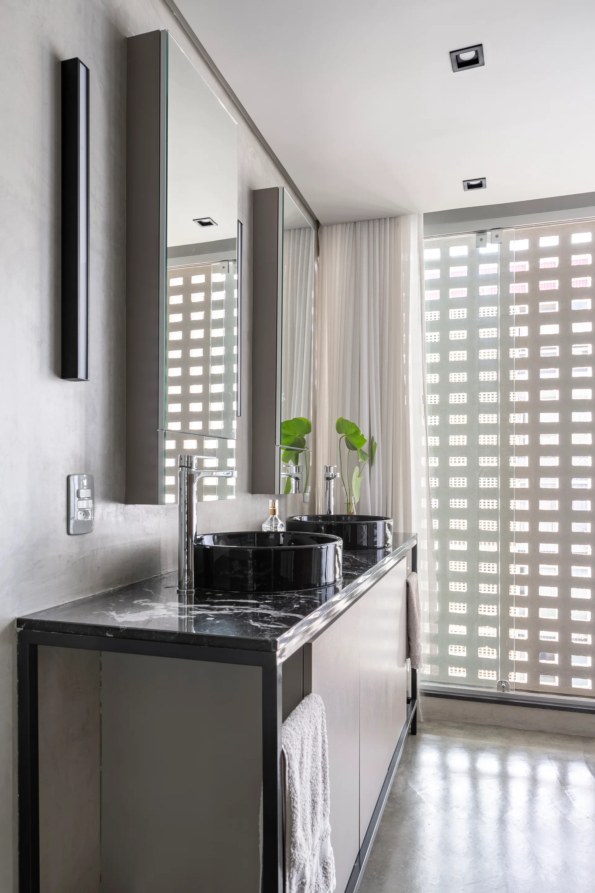 A modern bathroom features a double vanity with black marble countertops, round black vessel sinks, concrete walls, and a large grid-screen window.
