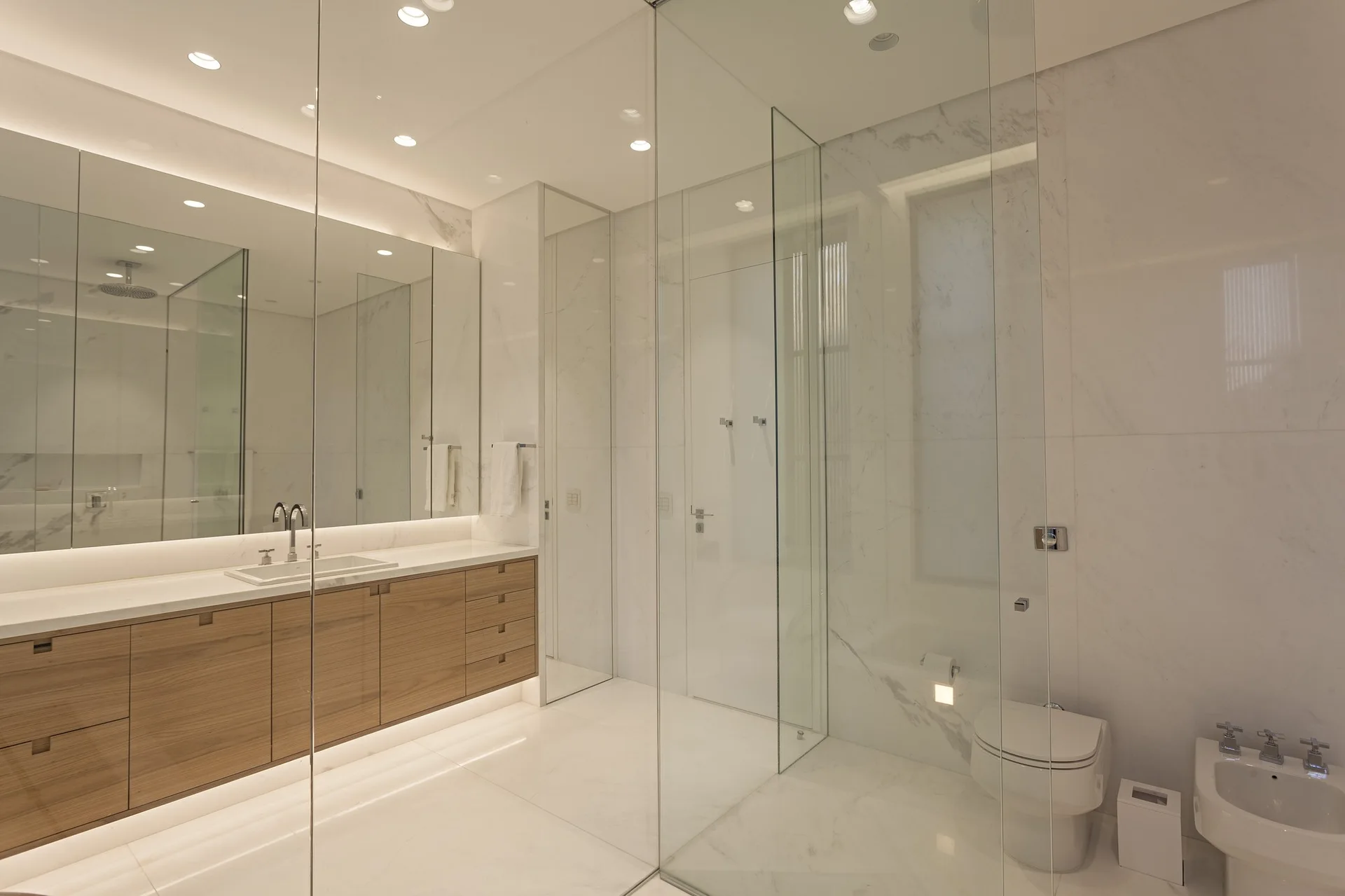 A minimalist bathroom features white marble tile walls, a long floating wood vanity with a large mirror, and seamless glass partitions.
