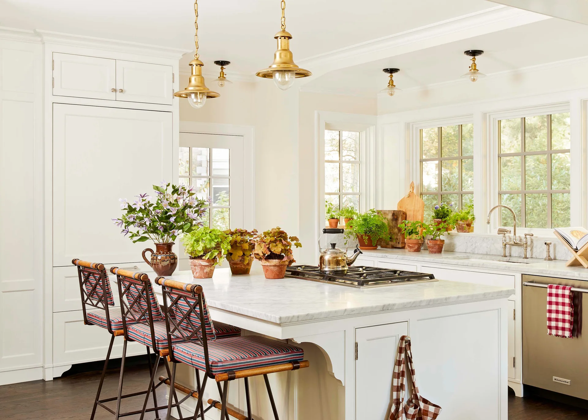 A white kitchen features a marble-topped island with striped stools, brass pendant lights, and potted plants near grid windows.