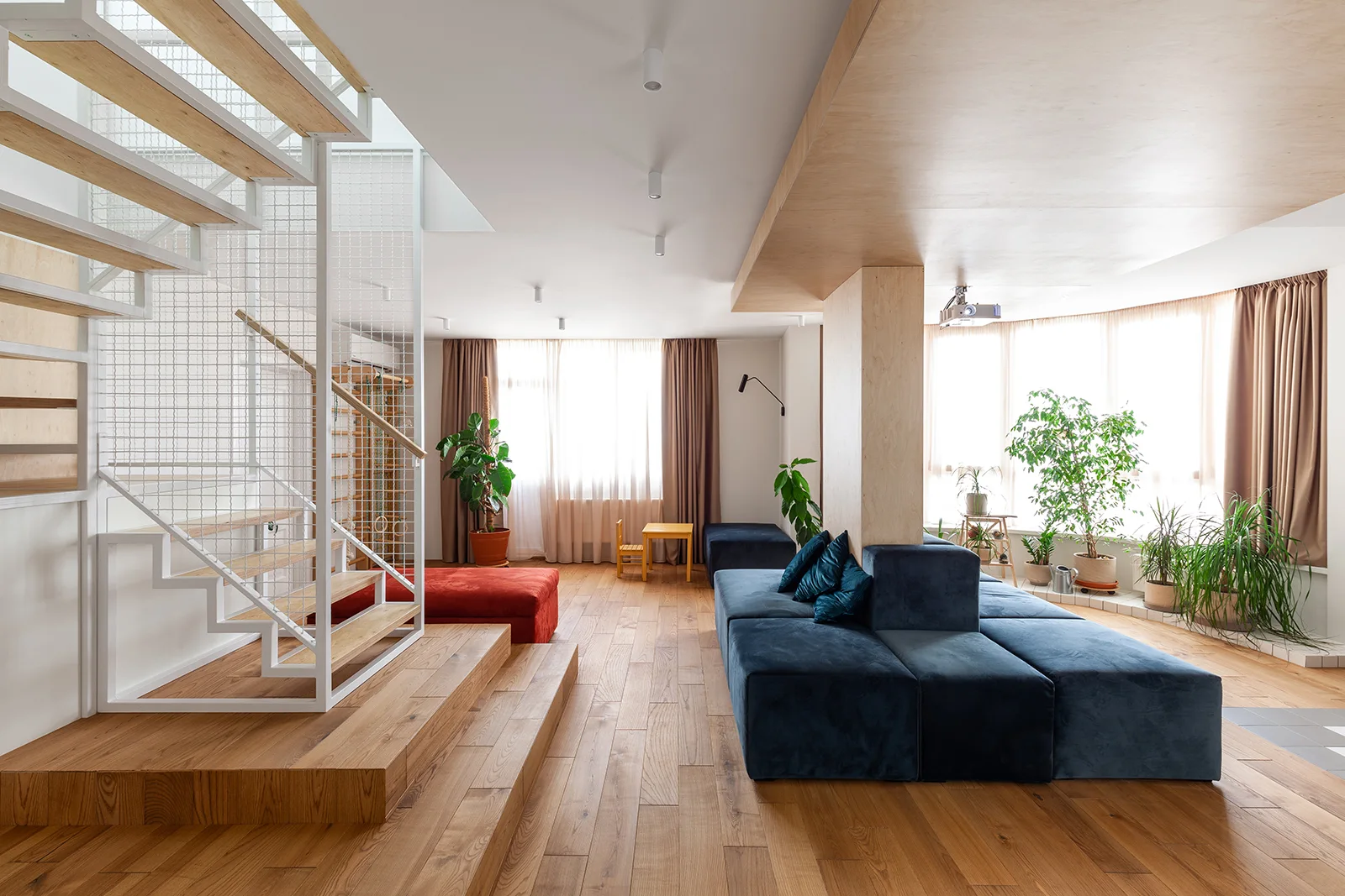 An open-plan living room with stepped wood flooring, a central modular blue velvet sofa, and an open-riser wood staircase.
